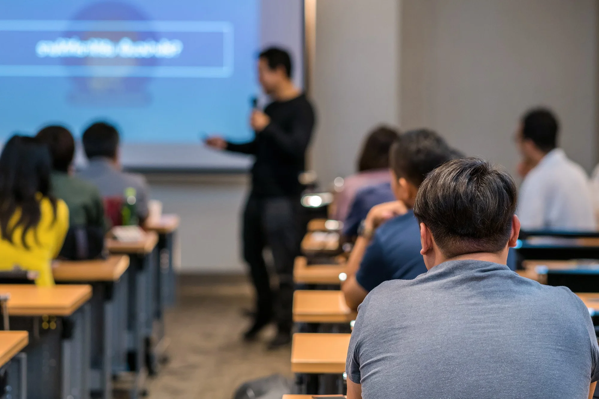 A blurred photo of a conference room with attendees sitting at desks, listening to a presenter standing near a large screen.