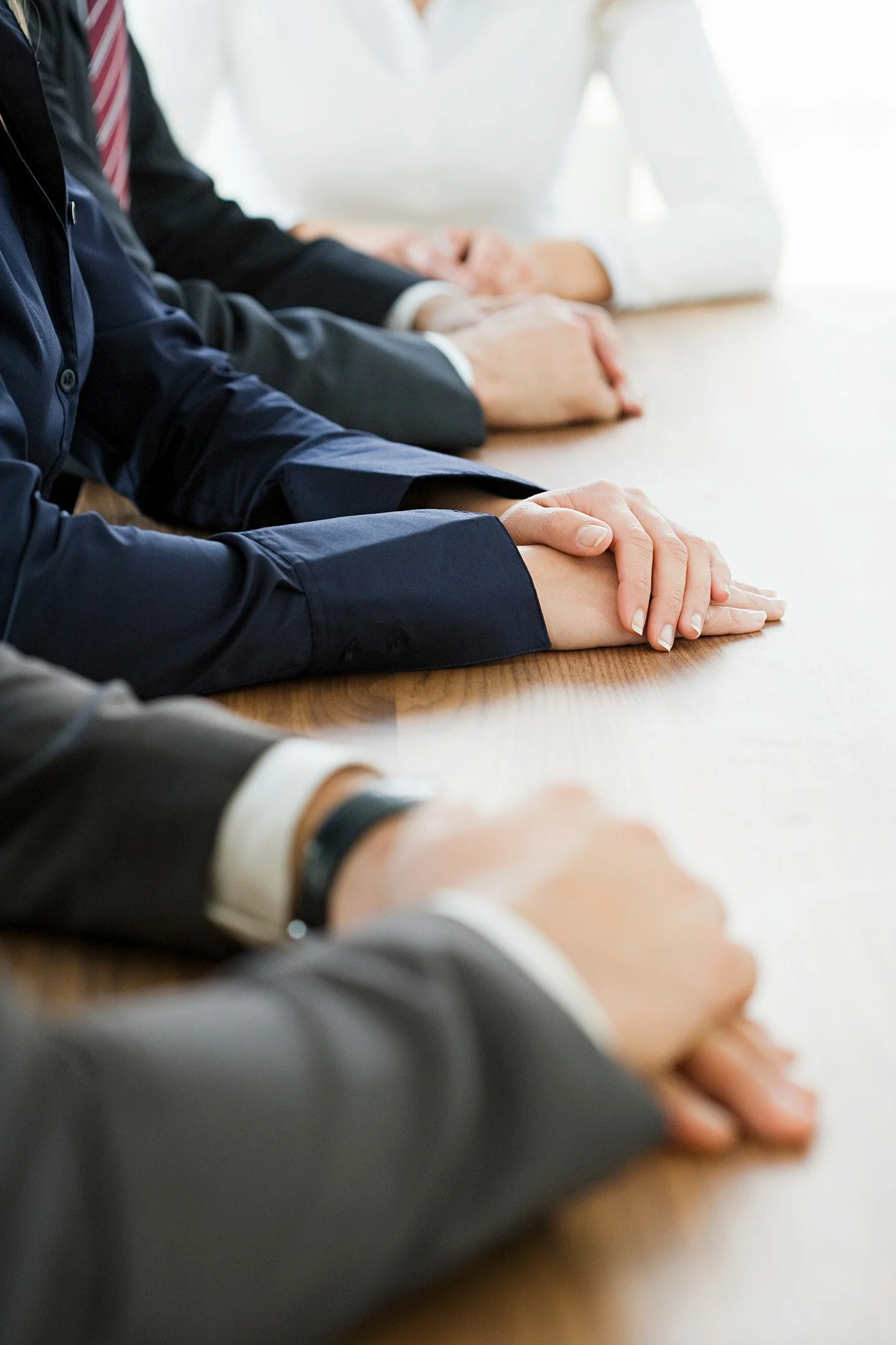 Close-up of people seated at a table with their hands clasped front and center, dressed in business attire, in a professional setting.