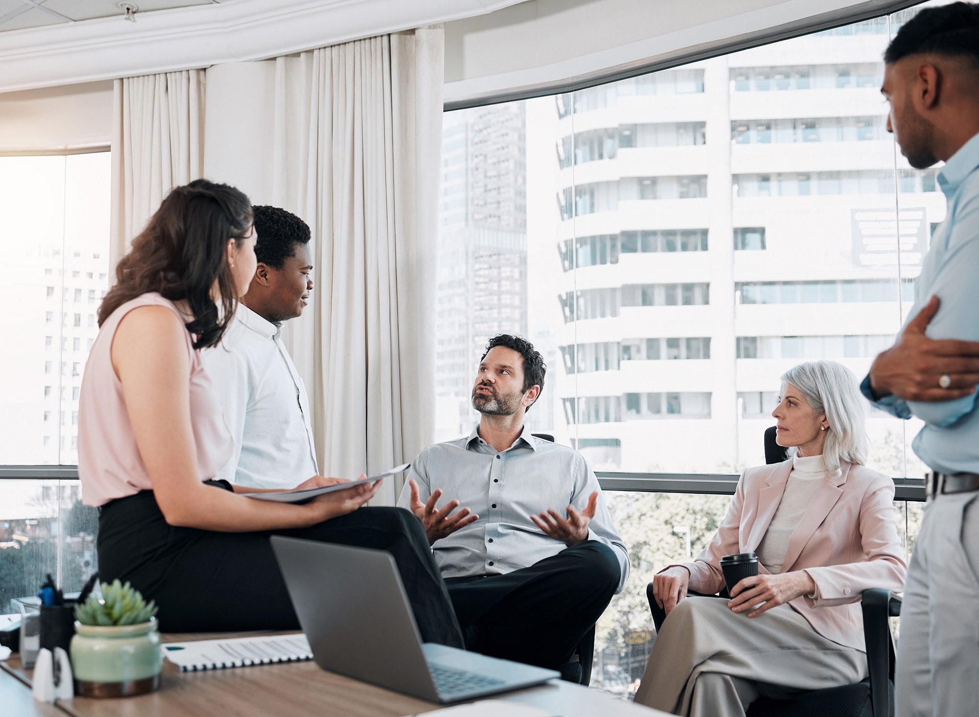 A diverse group of people in a modern office meeting room engaged in a discussion, with some standing and others seated, with large windows and city buildings outside.