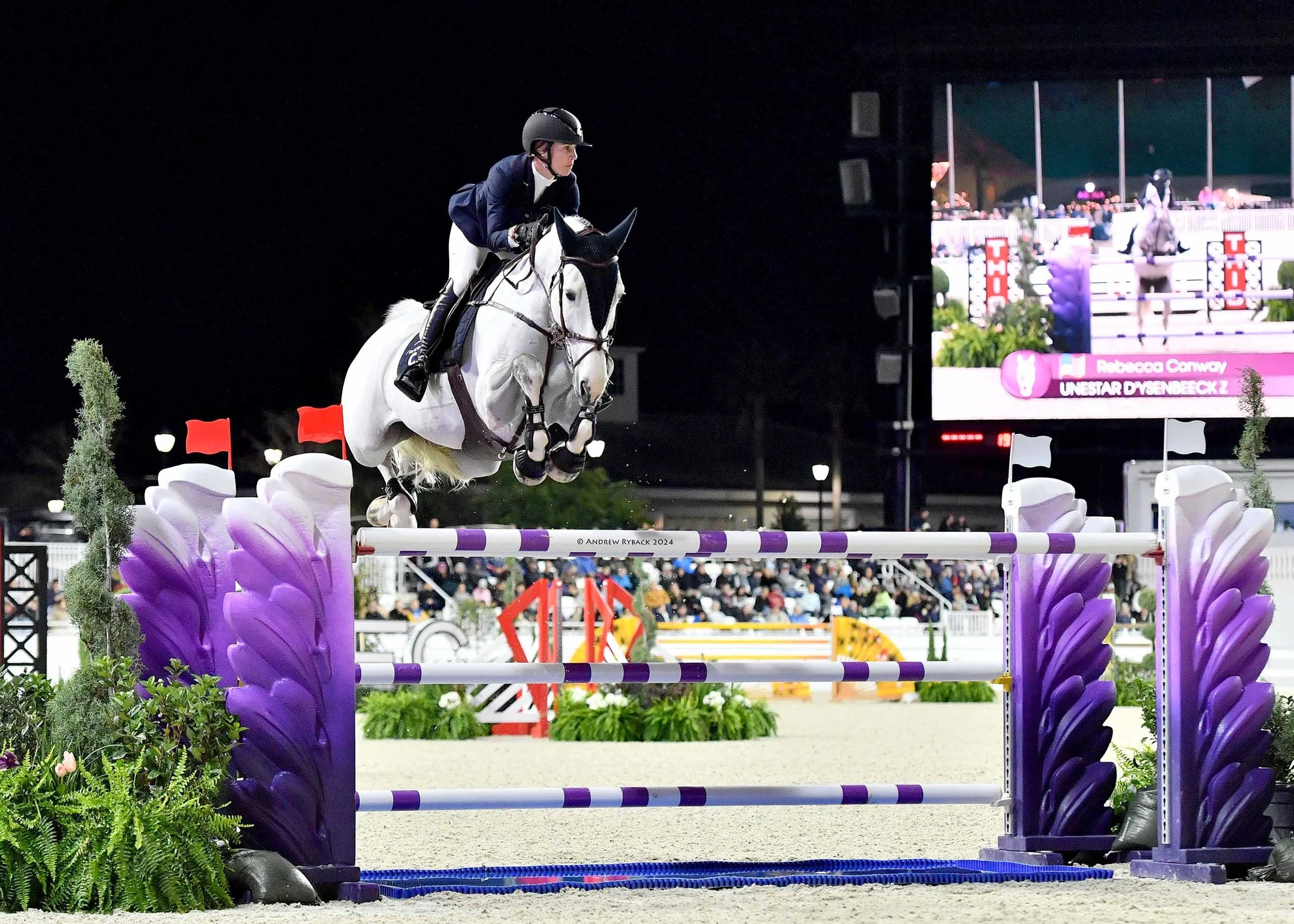 A rider in equestrian gear jumping over an obstacle on a white horse at night during a show jumping competition, with a digital scoreboard and audience in the background.