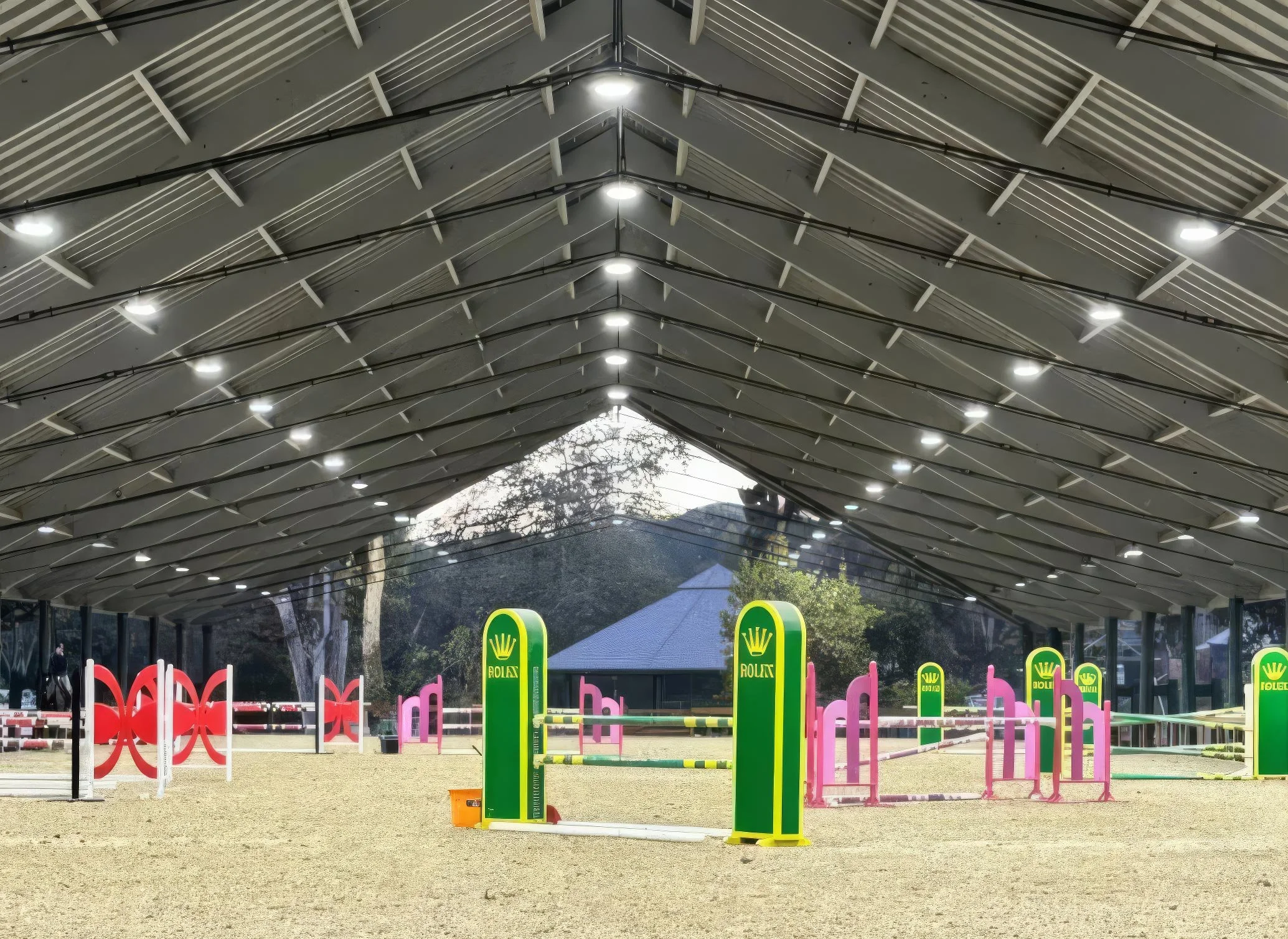 An outdoor horse jumping arena under a large, modern, metal roof structure with bright lights. The arena has colorful jumps, including green and pink poles and signs with the Rolex logo, and surrounding trees are visible in the background.