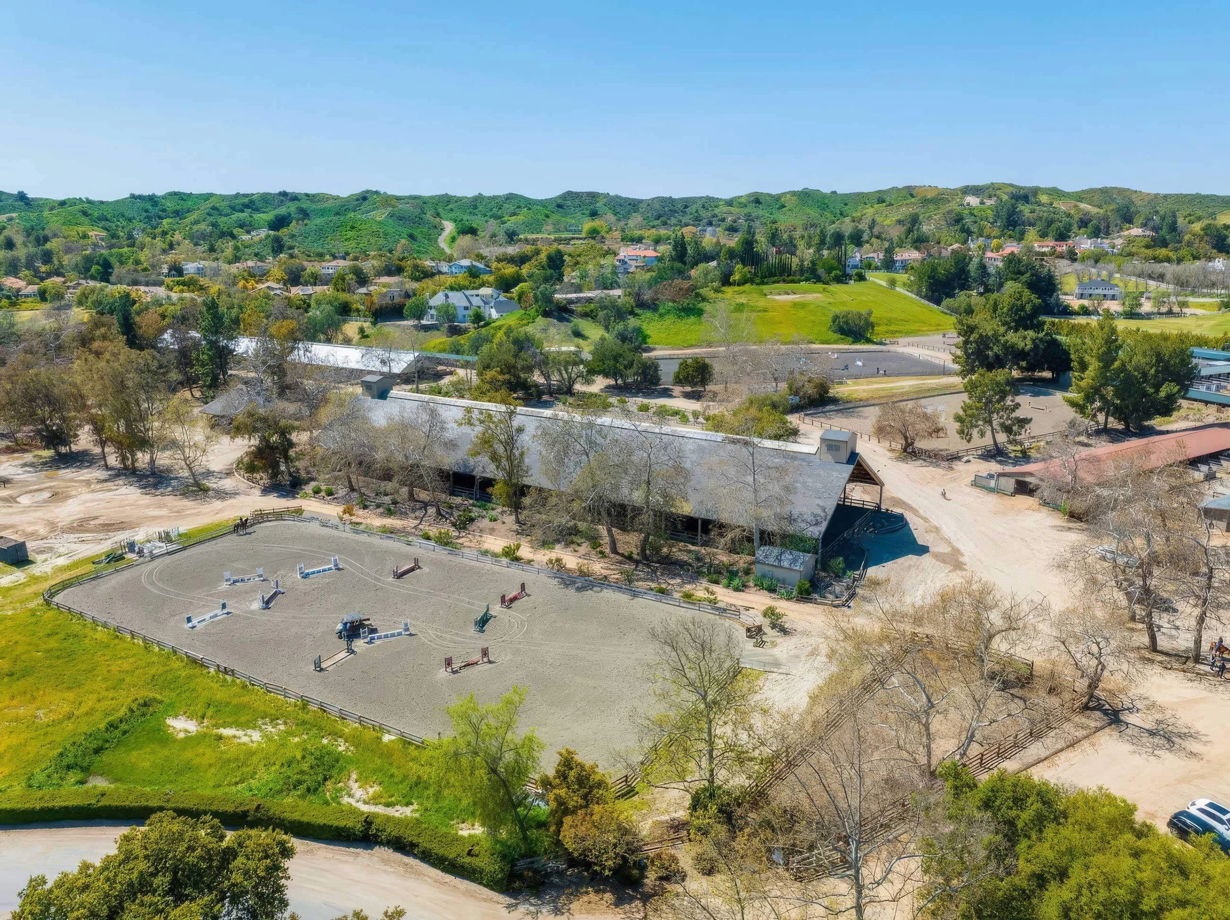 Aerial view of a grassy hill with houses, trees, and a dirt path in the background, and a horse riding ring with jumps and an arena in the foreground.