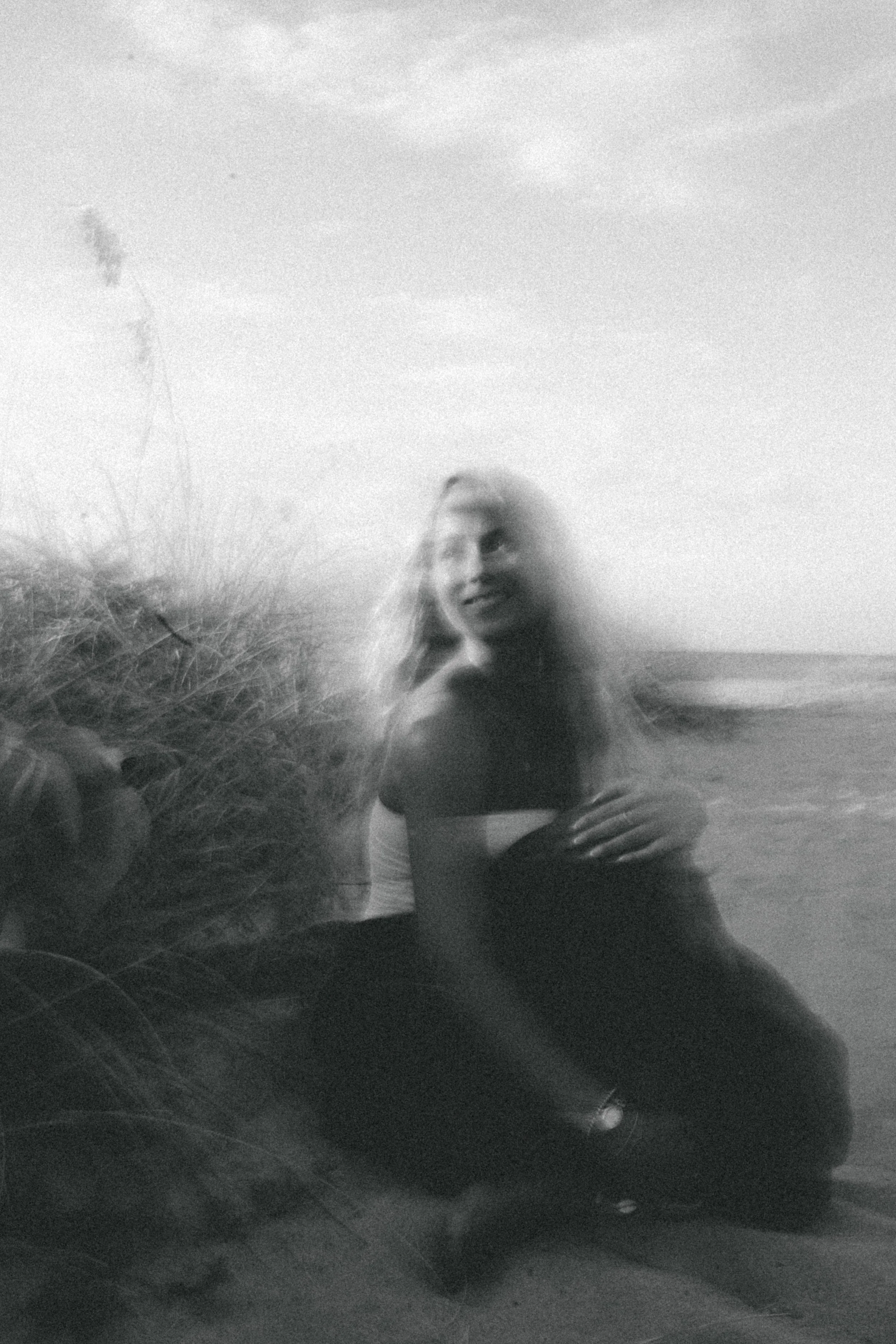 Blurry black-and-white photograph of a woman with long hair, sitting on the beach near dunes, smiling and looking away from the camera.