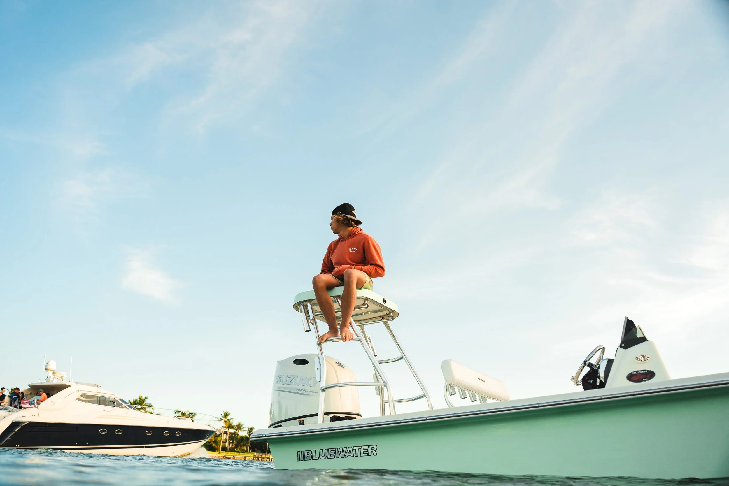 A person sitting on a boat's captain seat, wearing a cap and an orange hoodie, with a boat and another yacht in the background on a sunny day.