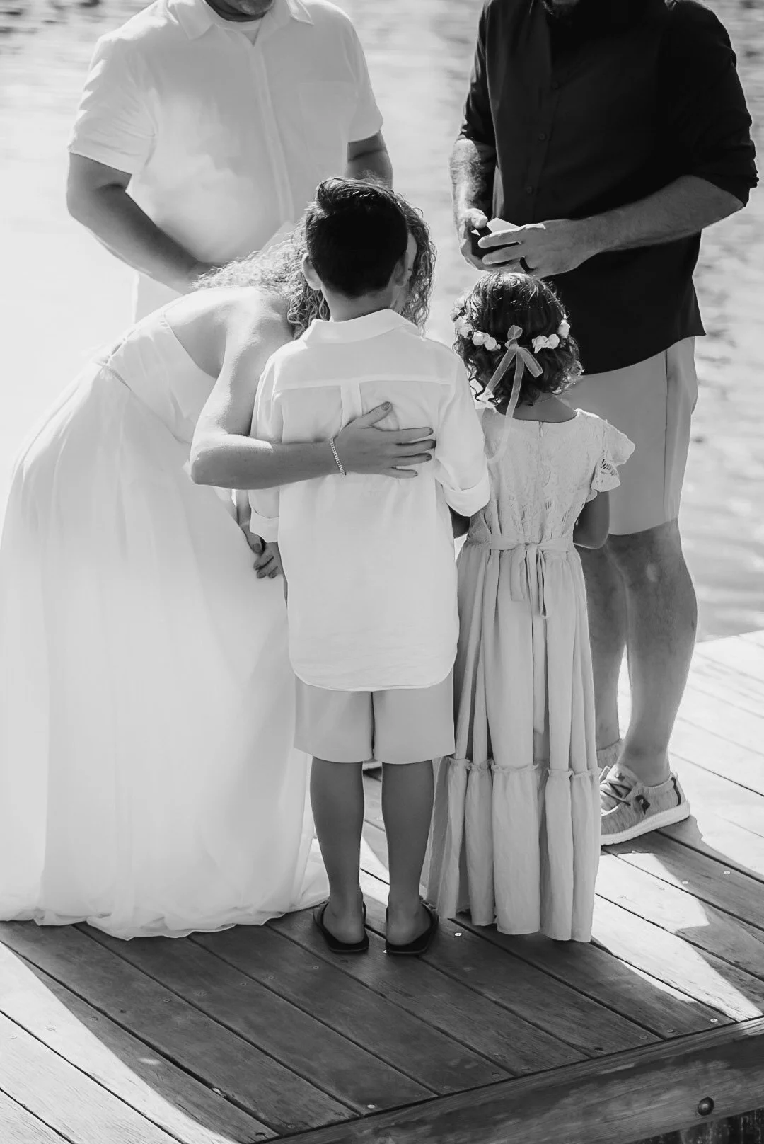 A group of people, including children and adults, gathered on a dock by the water, engaged in a ceremony, possibly a wedding or religious ritual.