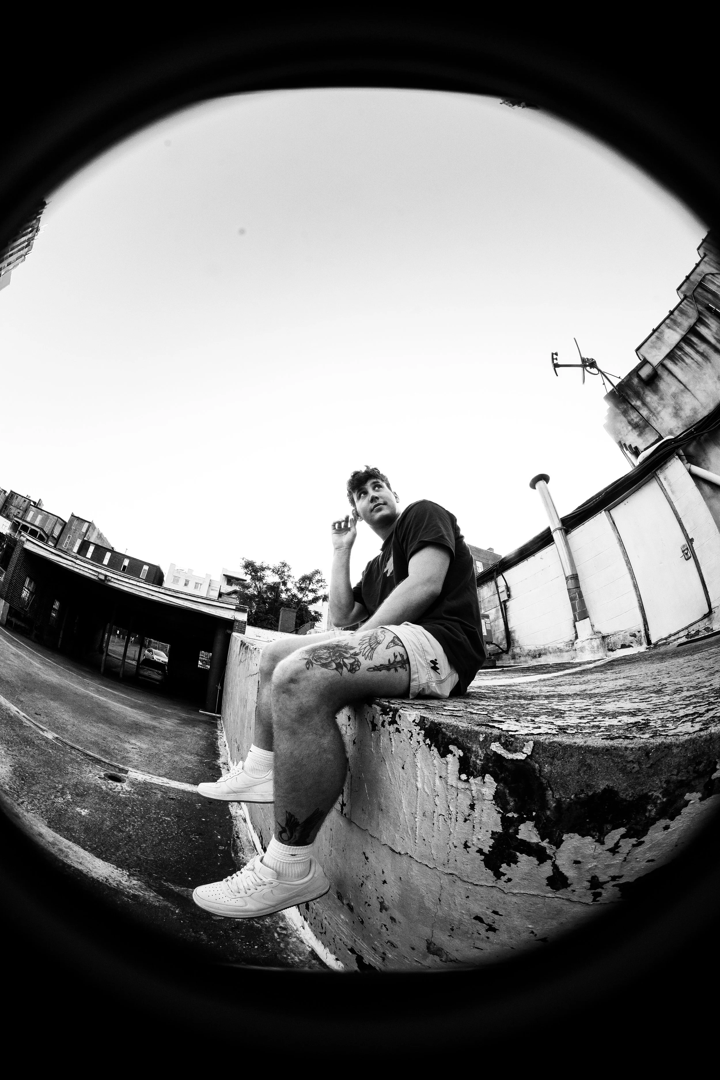 A young man with tattoos on his legs, wearing white sneakers, shorts, and a dark t-shirt, sitting on a weathered concrete ledge in an urban rooftop setting, looking away from the camera with a thoughtful expression.