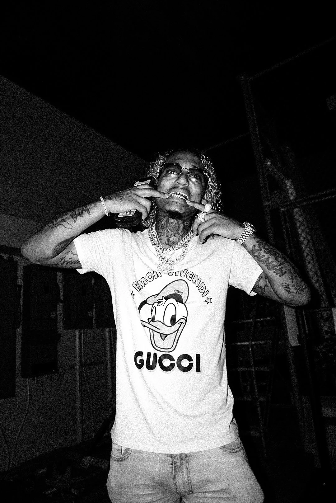 A young man with curly hair and tattoos, wearing glasses and a Gucci t-shirt with Donald Duck, is talking on a cell phone and biting his finger.