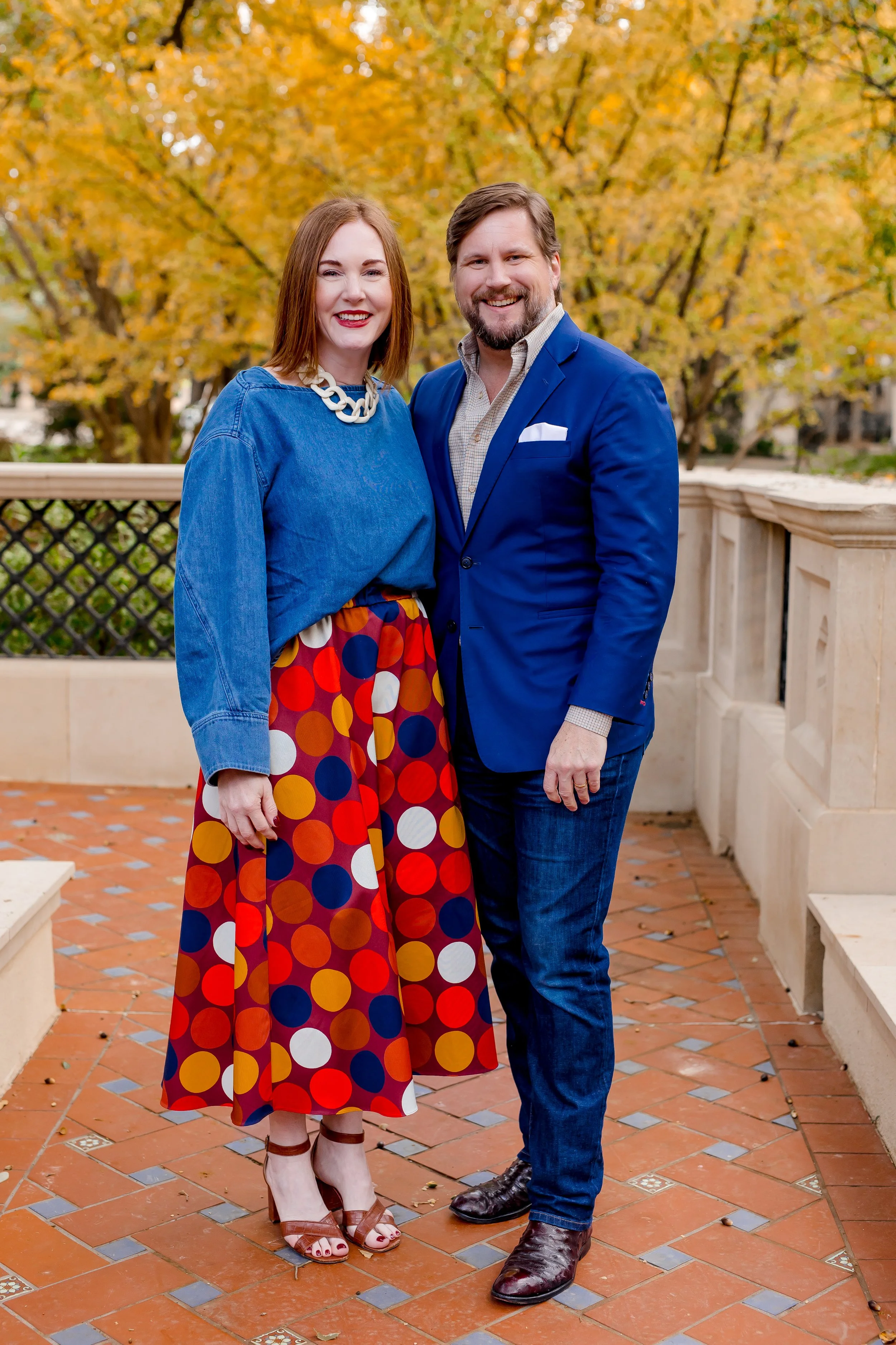 Two people posing together on a brick patio with a stone railing, dressed in colorful and formal-casual outfits, with trees displaying yellow fall foliage in the background.