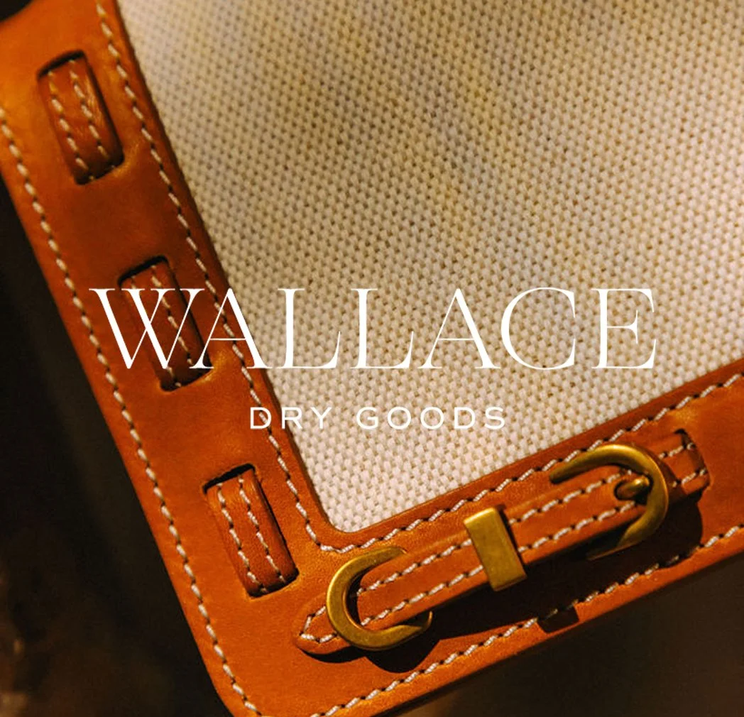 Close-up of a tan leather and canvas bag with stitched detailing and a brass buckle, featuring the text “WALLACE DRY GOODS” centered on the fabric panel.
