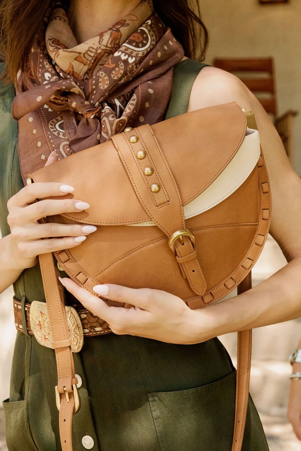 Close-up of hands holding a structured brown leather crossbody bag with a flap closure, brass hardware, and decorative stitching, paired with a patterned scarf and belt.
