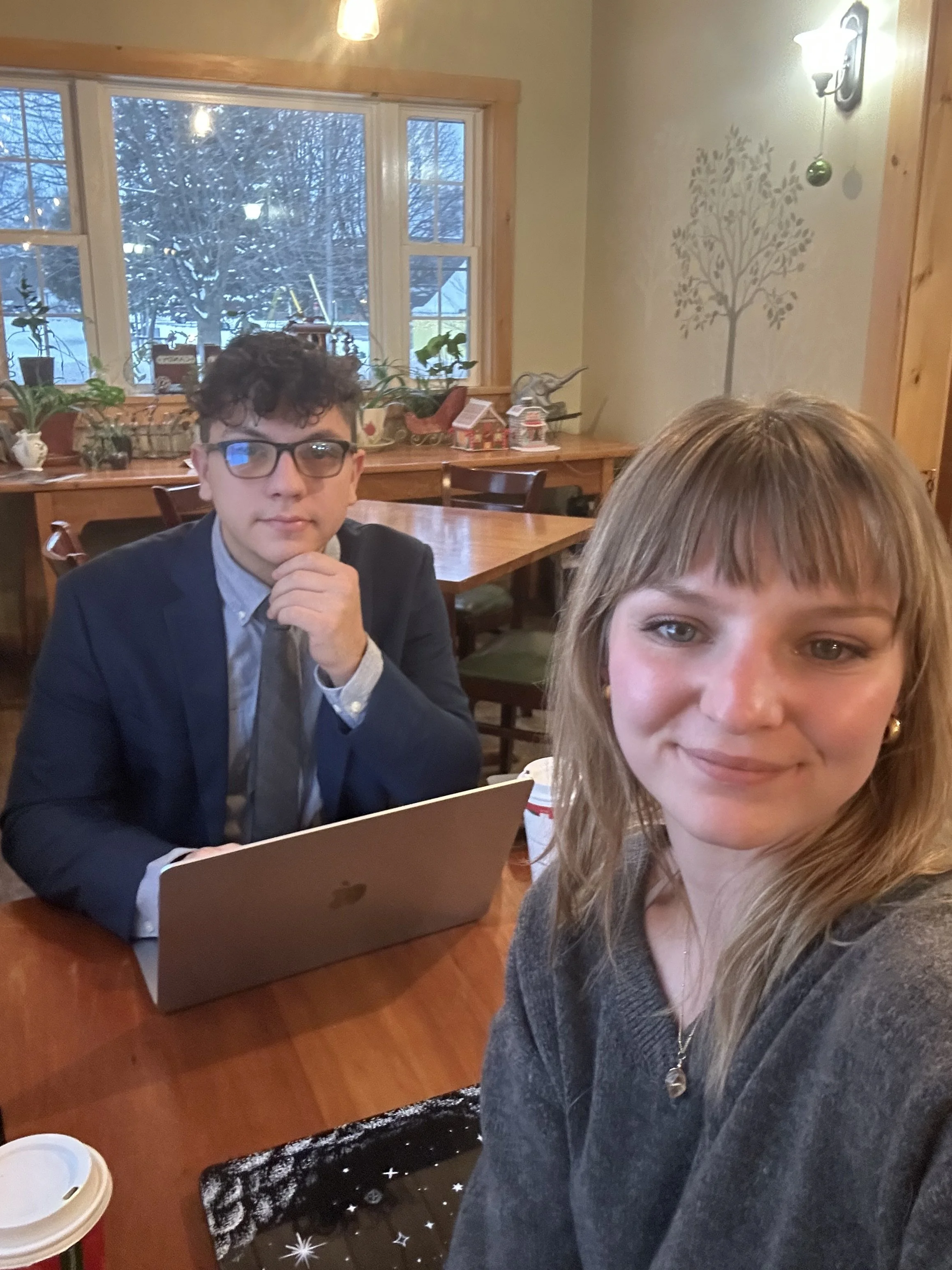 A woman taking a selfie in a room with wooden decor, next to a young man wearing glasses, sitting at a table with a laptop. The background shows a window with a snowy outdoor scene and decorative plants.