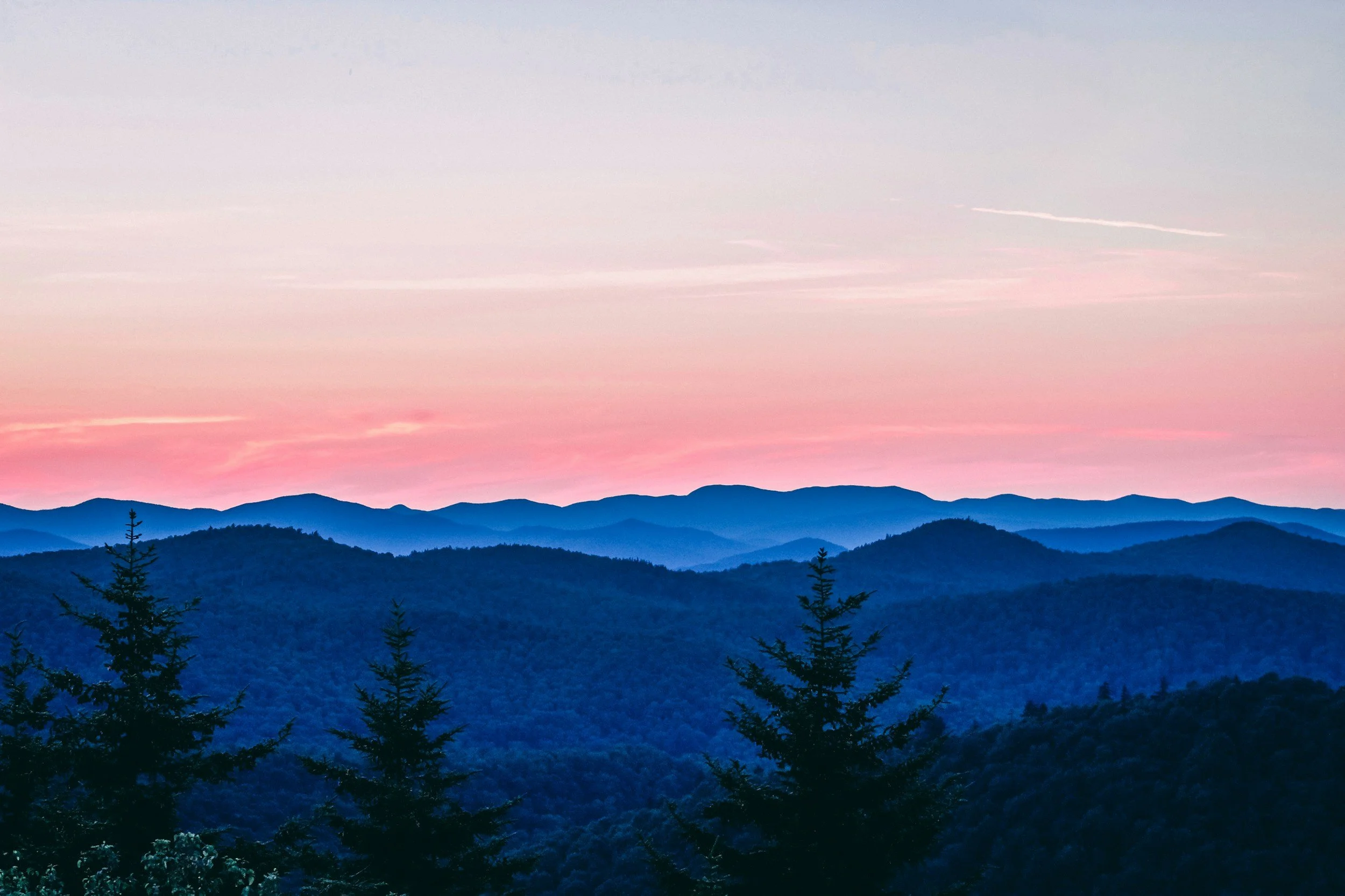 Scenic landscape of blue mountains under a pastel pink and purple sky at dawn or dusk, with silhouette of trees in the foreground.