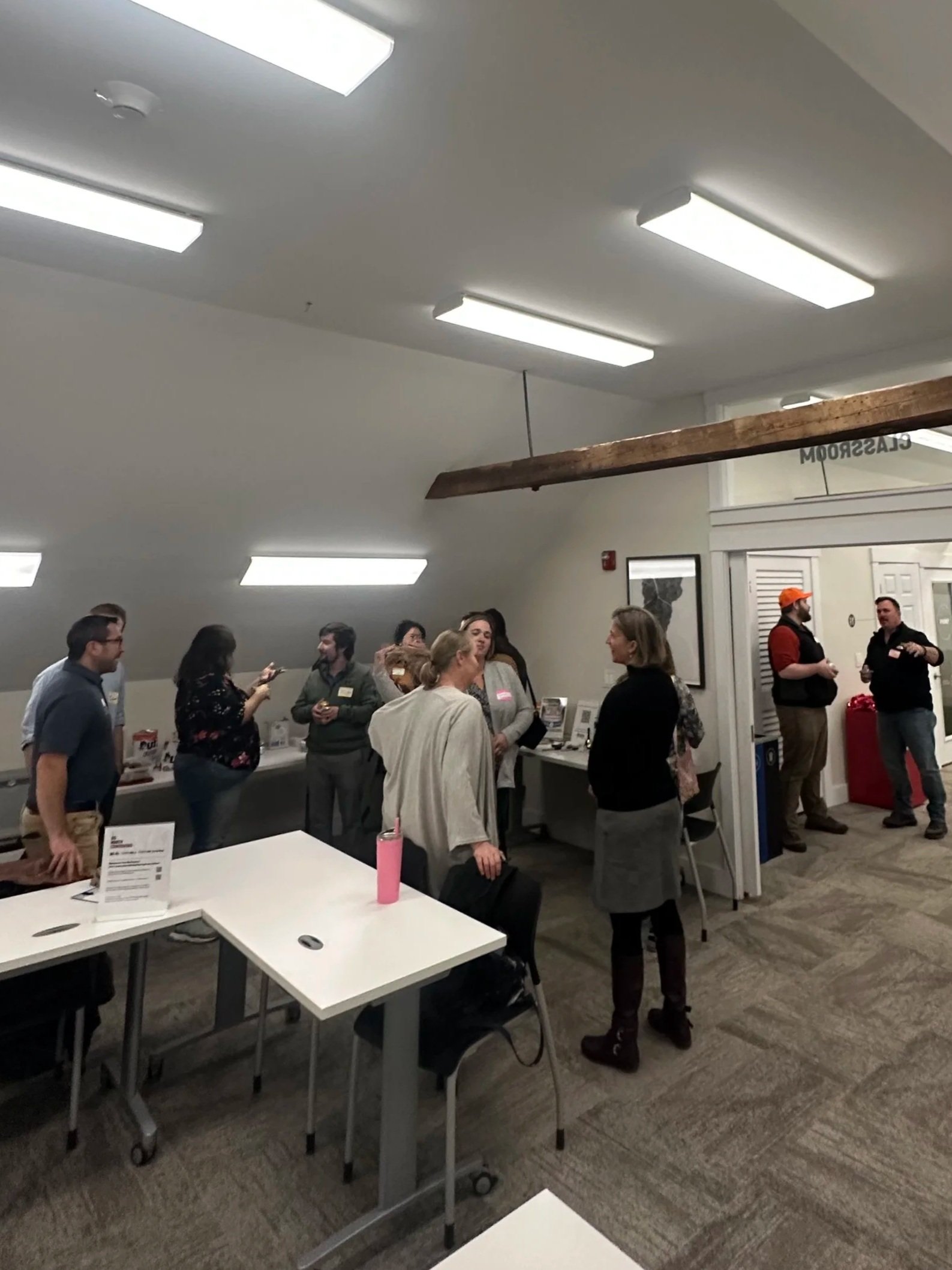 People gathered in a room, standing in groups, chatting, with tables and a bright ceiling light.