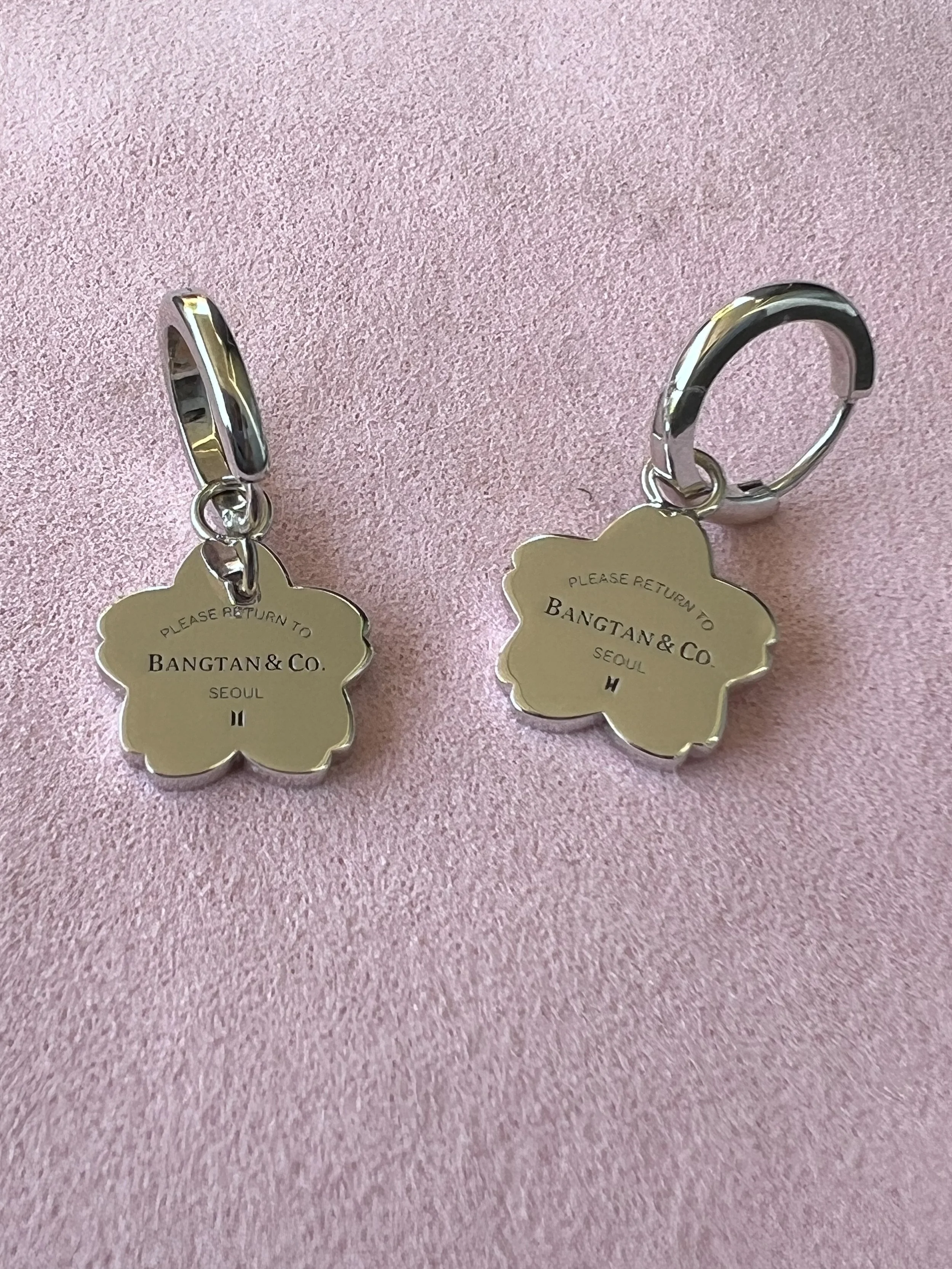 Two silver earrings with flower-shaped charms engraved with "Please return to Bangtan & Co. Seoul" and different initials, "H" and "II", on a pink textured background.