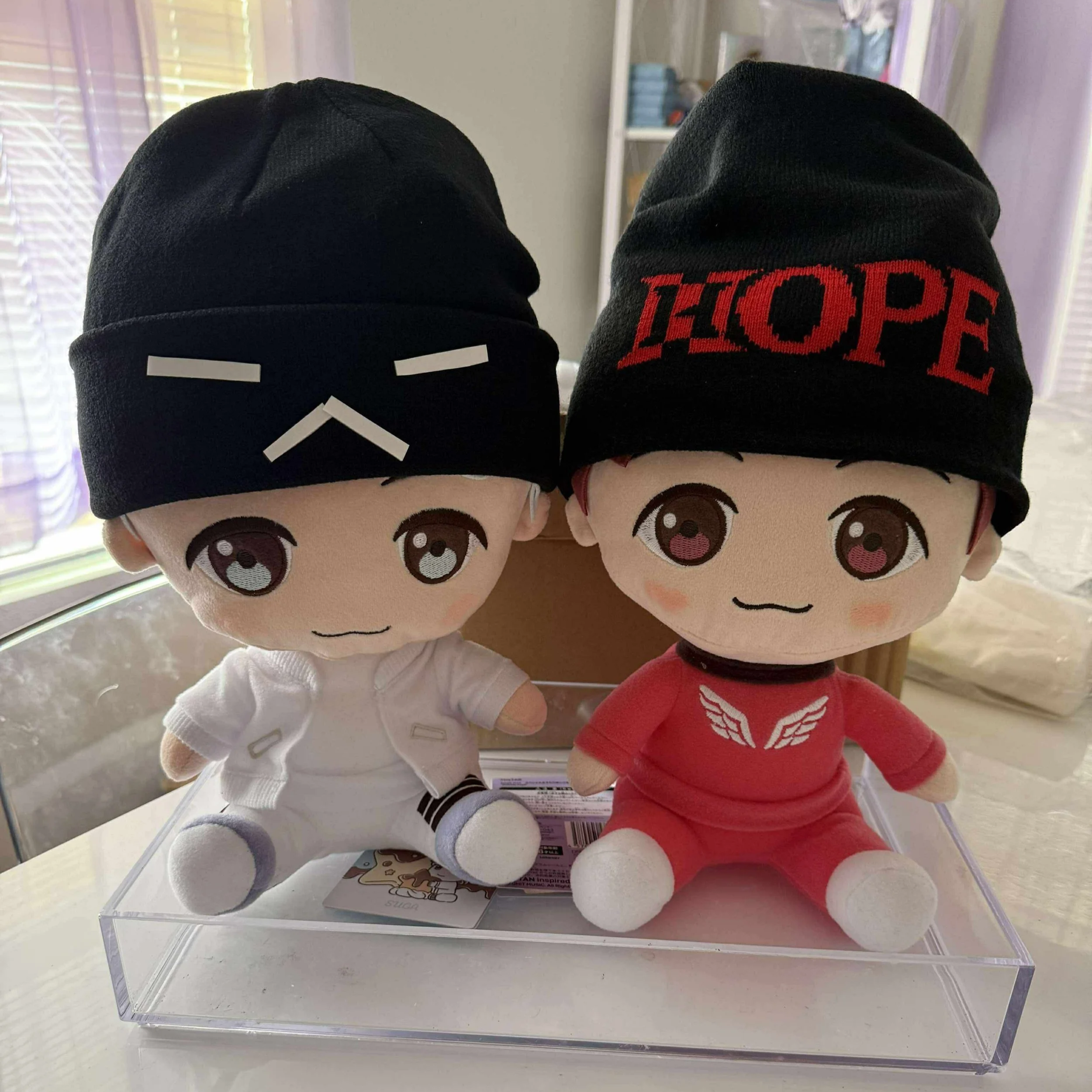 Two plush dolls with large eyes, wearing black beanie hats. The doll on the left is dressed in white, and the doll on the right is dressed in red with the word "HOPE" on the hat.