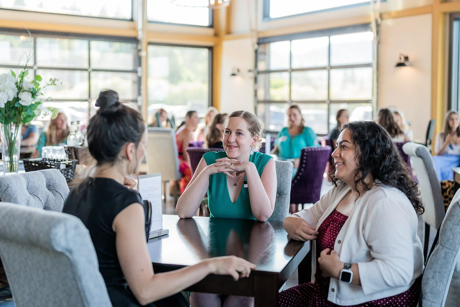 Three women are sitting at a table in a local Southern Oregon winery, engaged in conversation, with several other women seated at tables in the background.