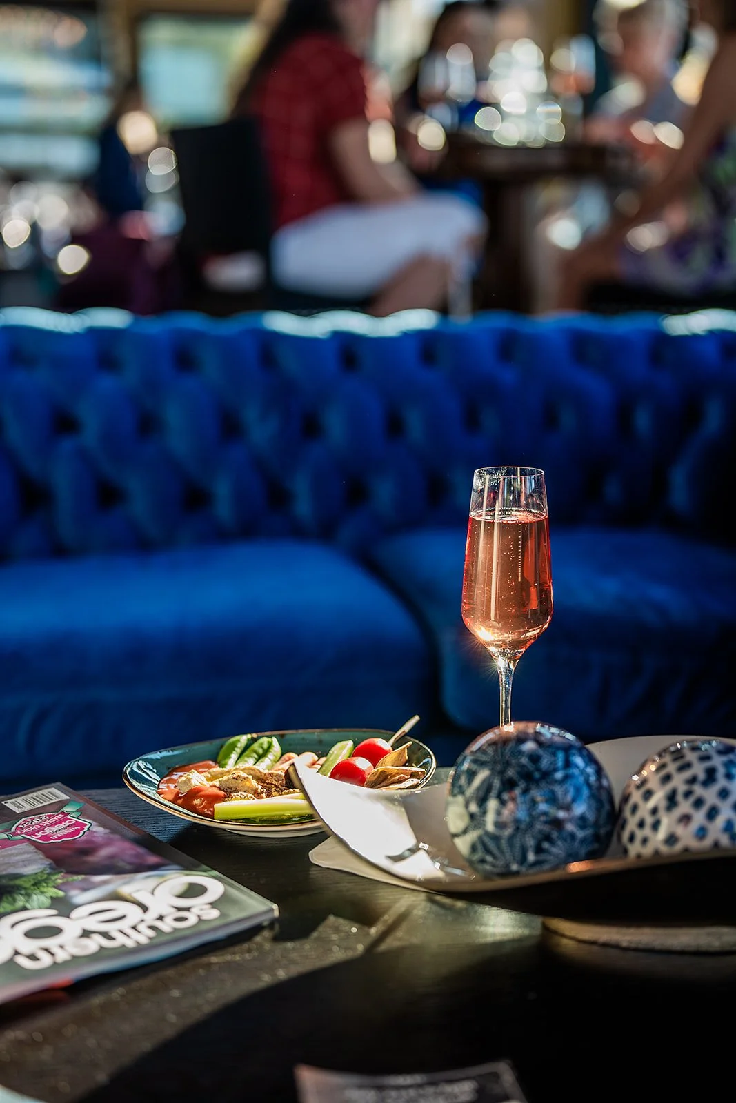 A table with a plate of vegetables and a glass of rosé wine, with blurred people sitting in the background.