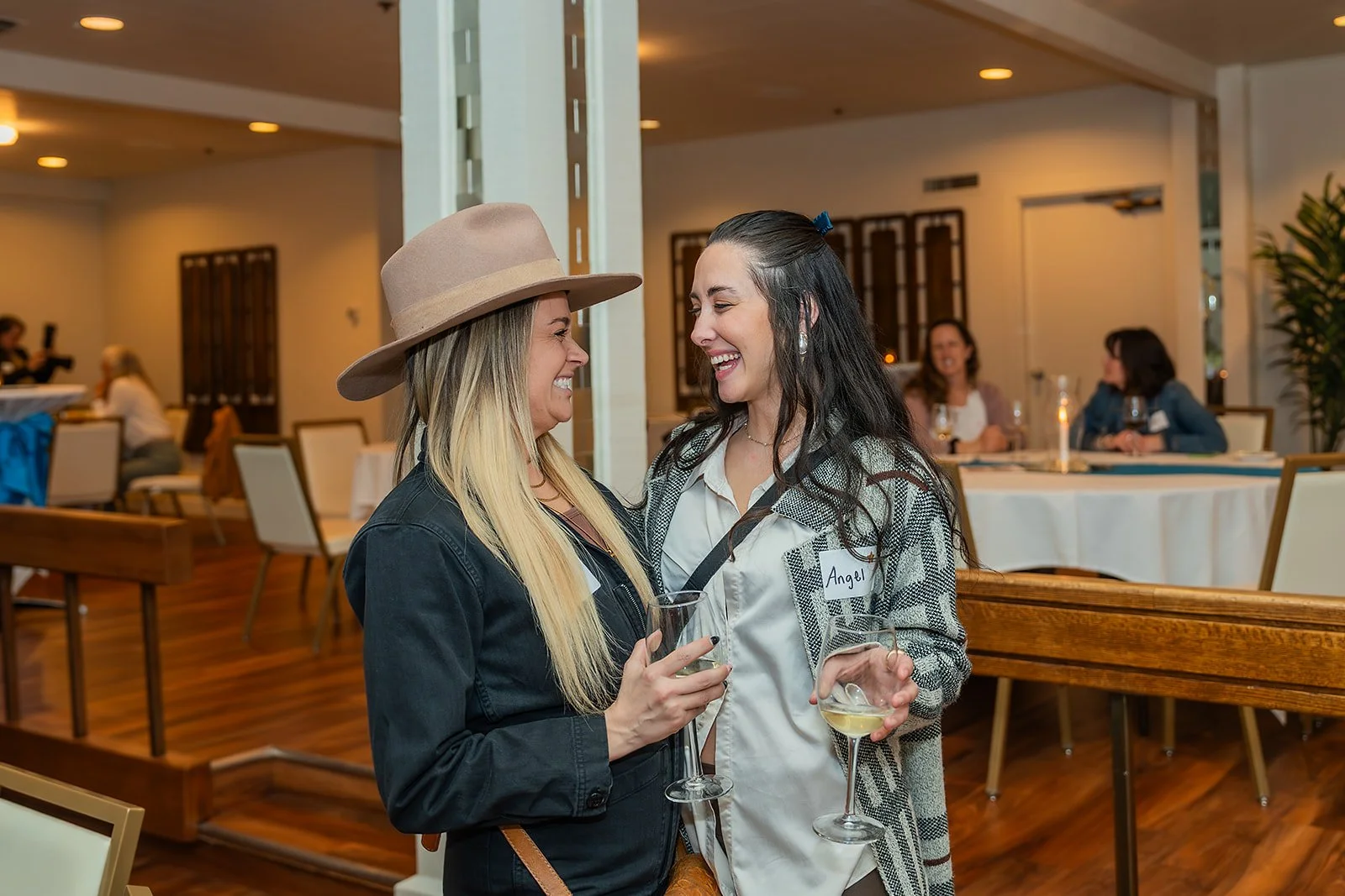Two women laughing at a Rogue Valley Women in Business event in Medford, OR with other guests seated at tables in the background.