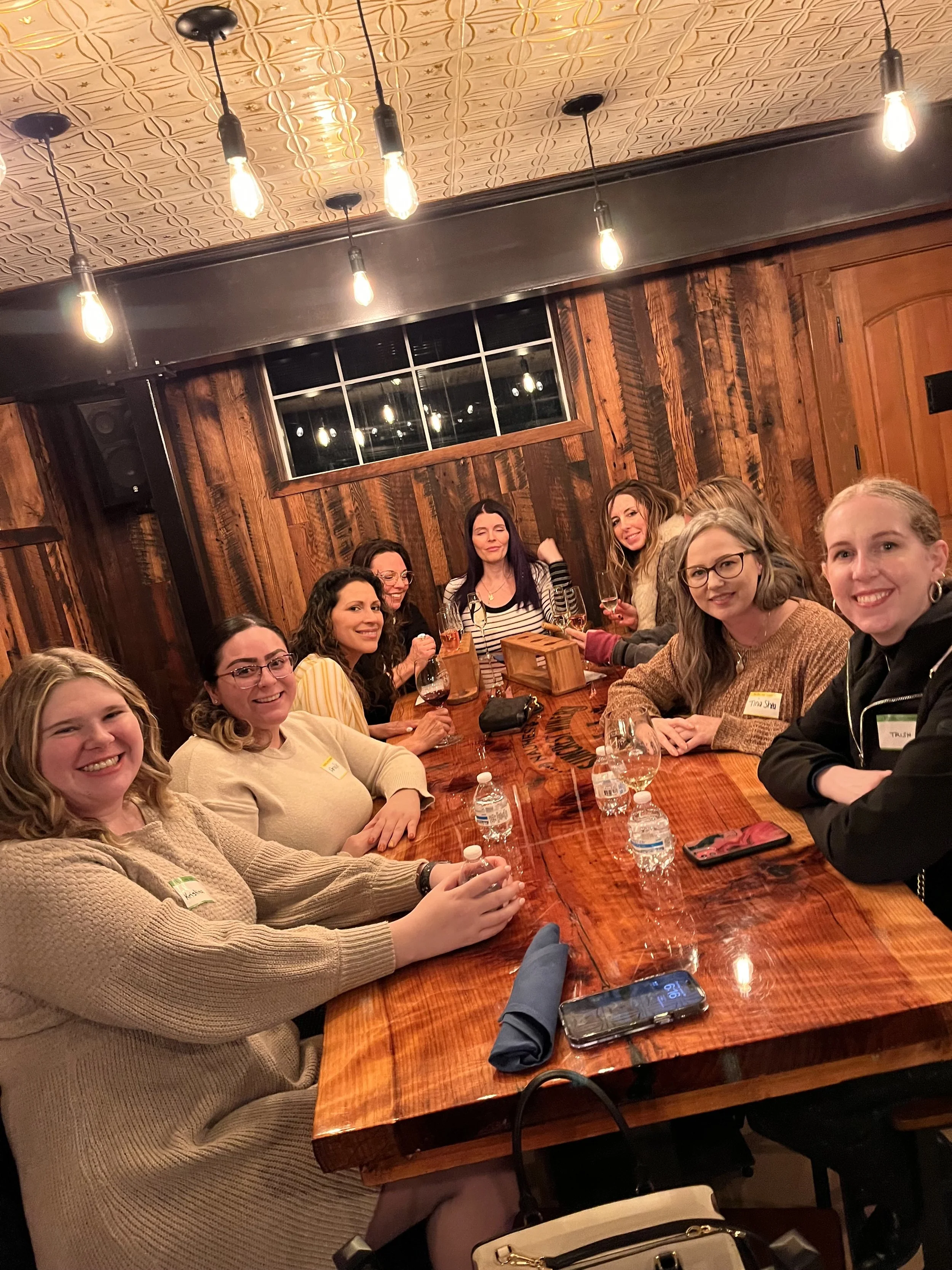 A group of nine women sitting around a wooden table in a cozy, rustic restaurant, smiling and holding drinks in Medford, OR.