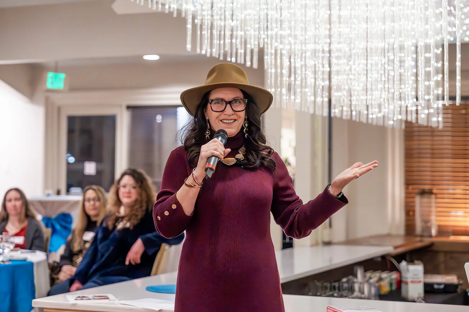 A woman wearing a wide-brimmed hat, glasses, and a maroon dress is speaking into a microphone at a Rogue Valley Women in Business event.