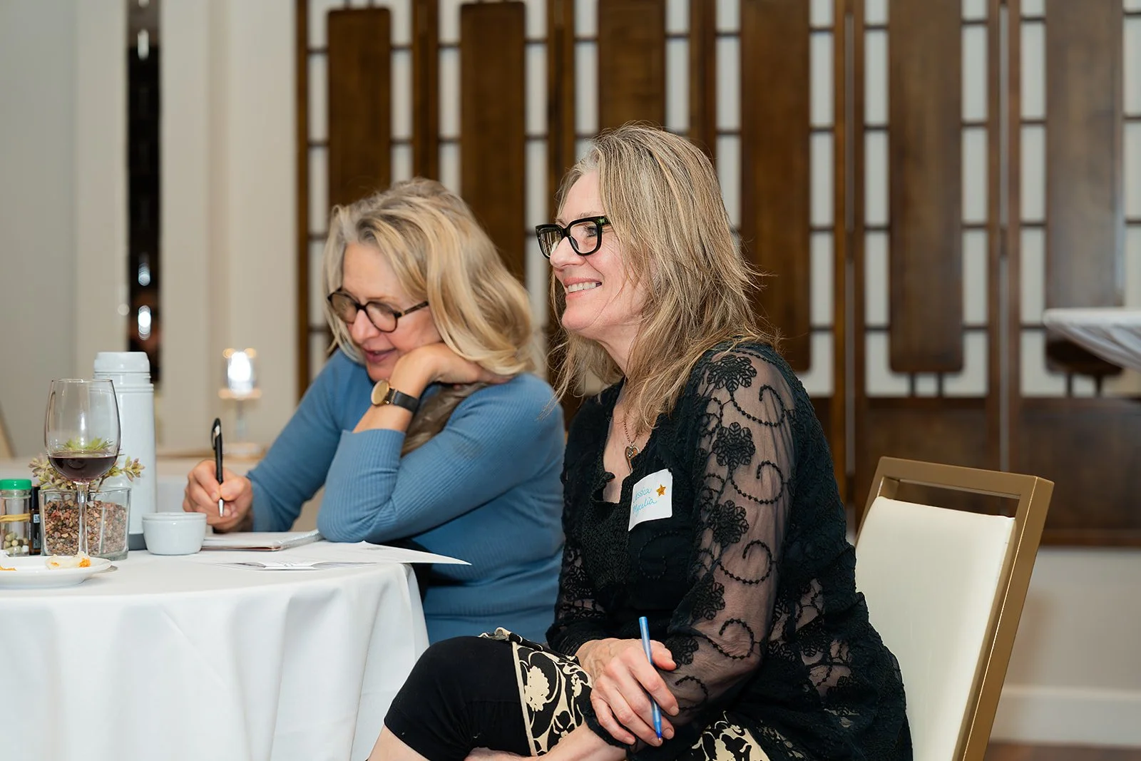 Two women sitting at a table during a Rogue Valley Women in Business mixer, one with a notepad and pen, smiling and looking to the side, with a glass of red wine.
