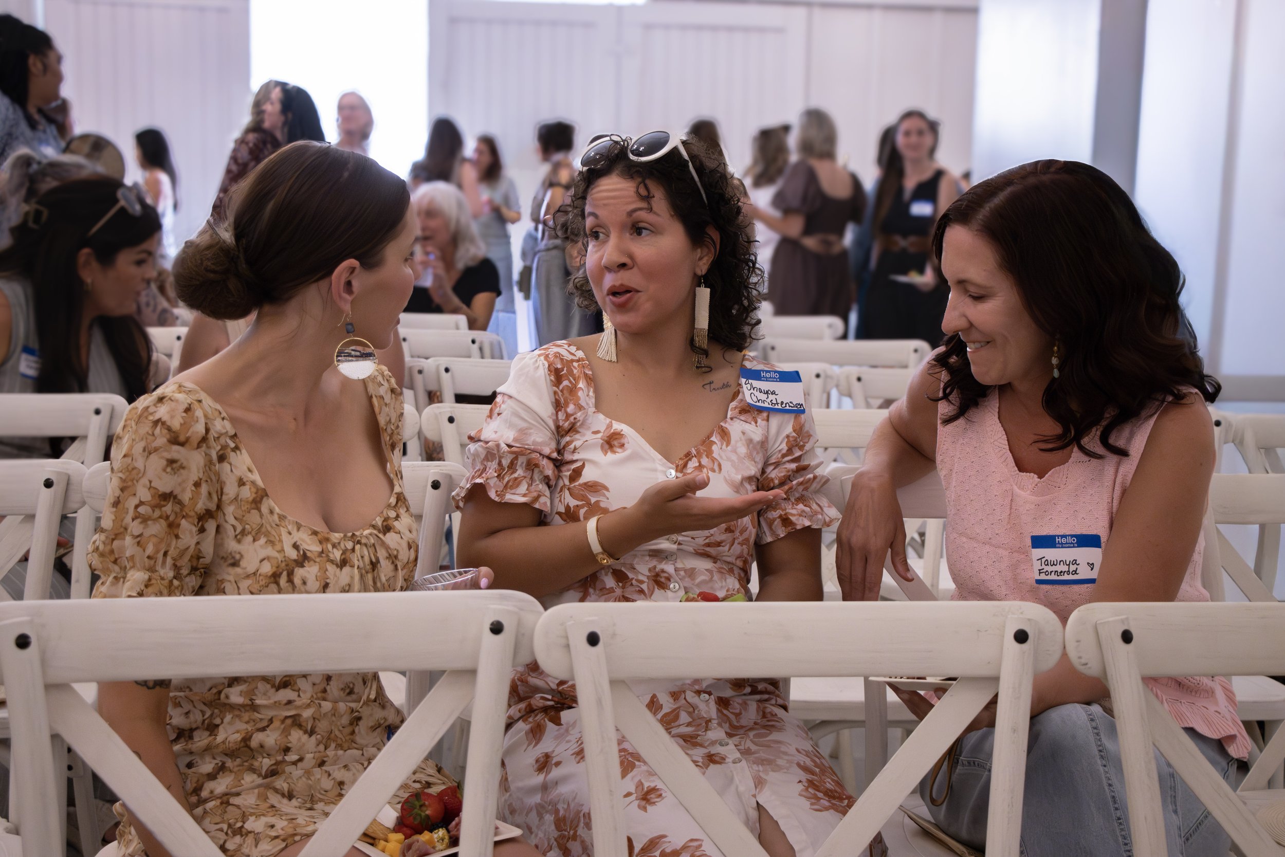 Three women are sitting at a table in a local Southern Oregon winery, engaged in conversation, with several other women seated at tables in the background.