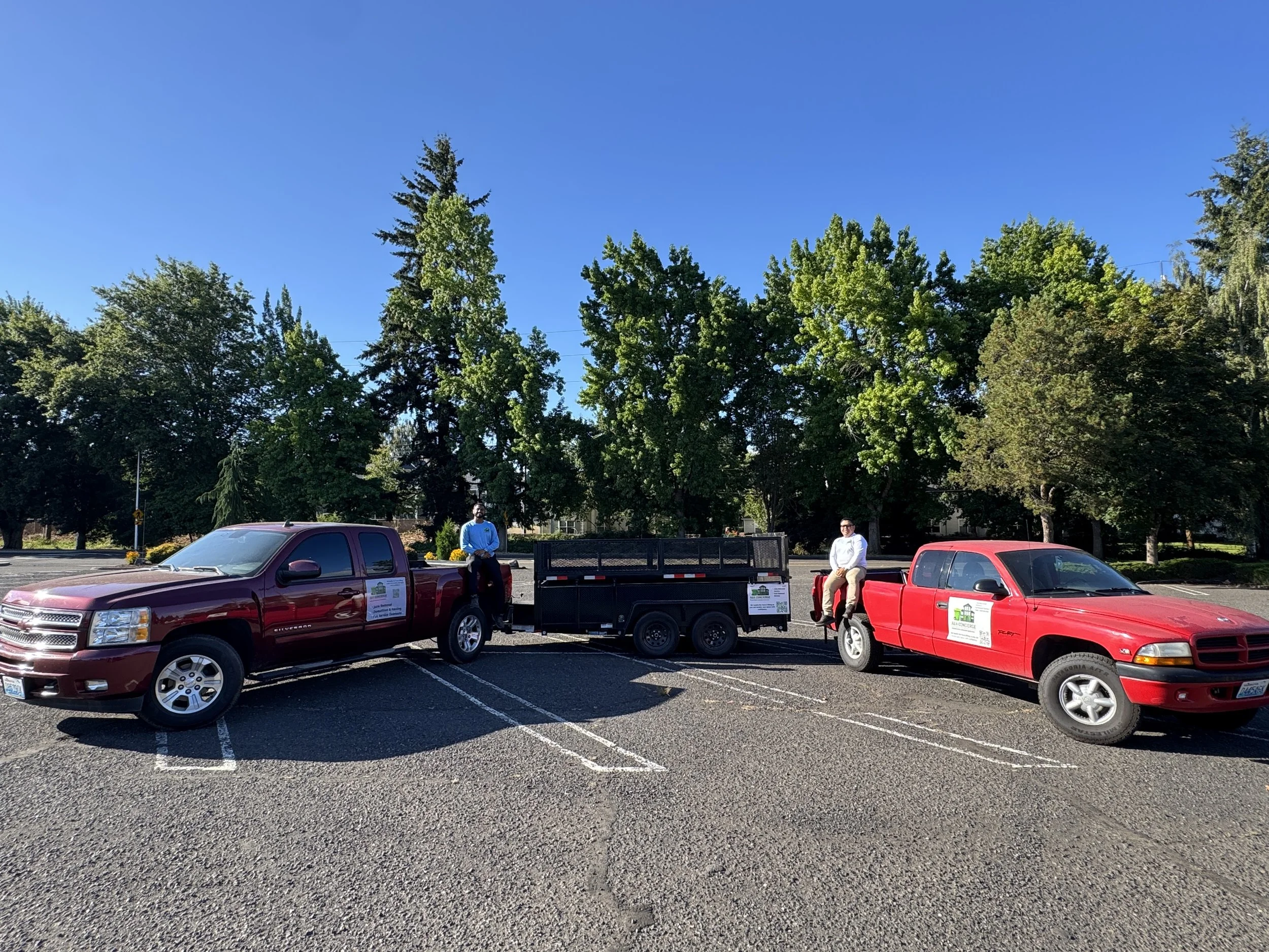 A parking lot with several cars, two men sitting on the edge of a red truck, and trees with green leaves under a clear blue sky.