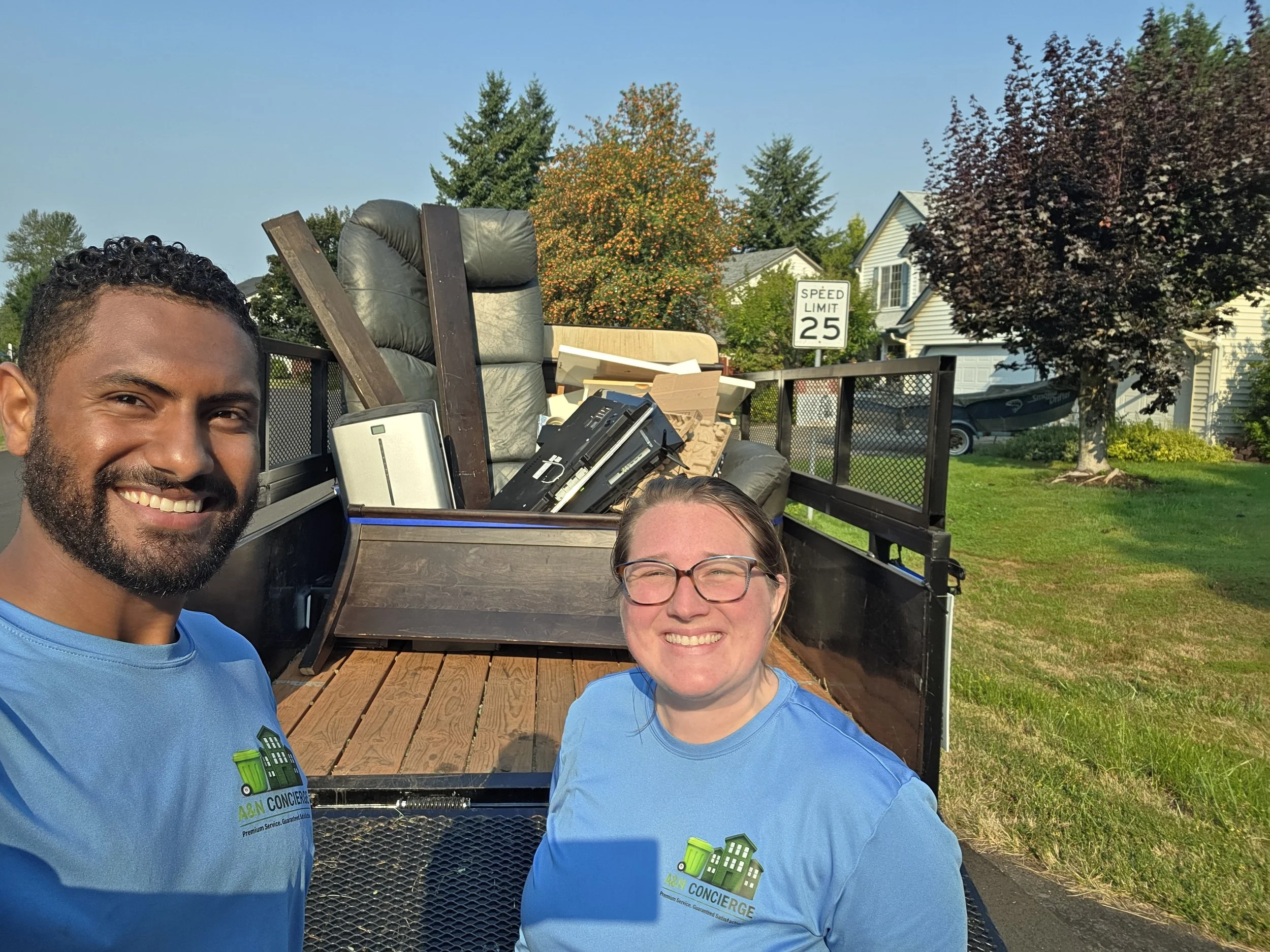 Two smiling individuals wearing blue shirts with logo, standing outdoors in front of a trailer filled with furniture and electronic items, with trees, houses, and a speed limit sign in the background.