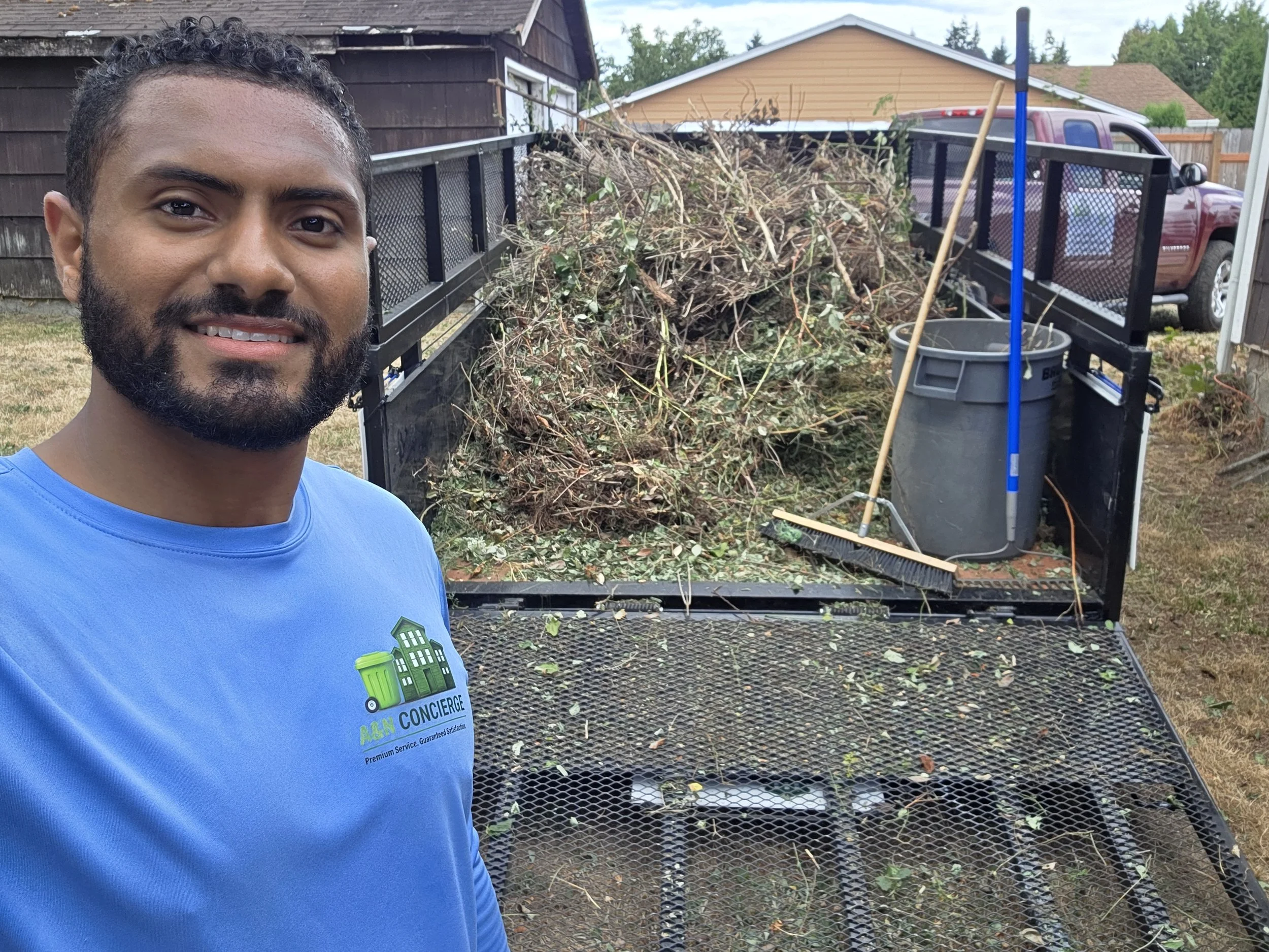 A man smiling in a blue shirt standing in front of a flatbed trailer filled with garden debris such as branches and leaves. The trailer is parked outdoors next to a yard with a brown wooden fence and houses in the background.