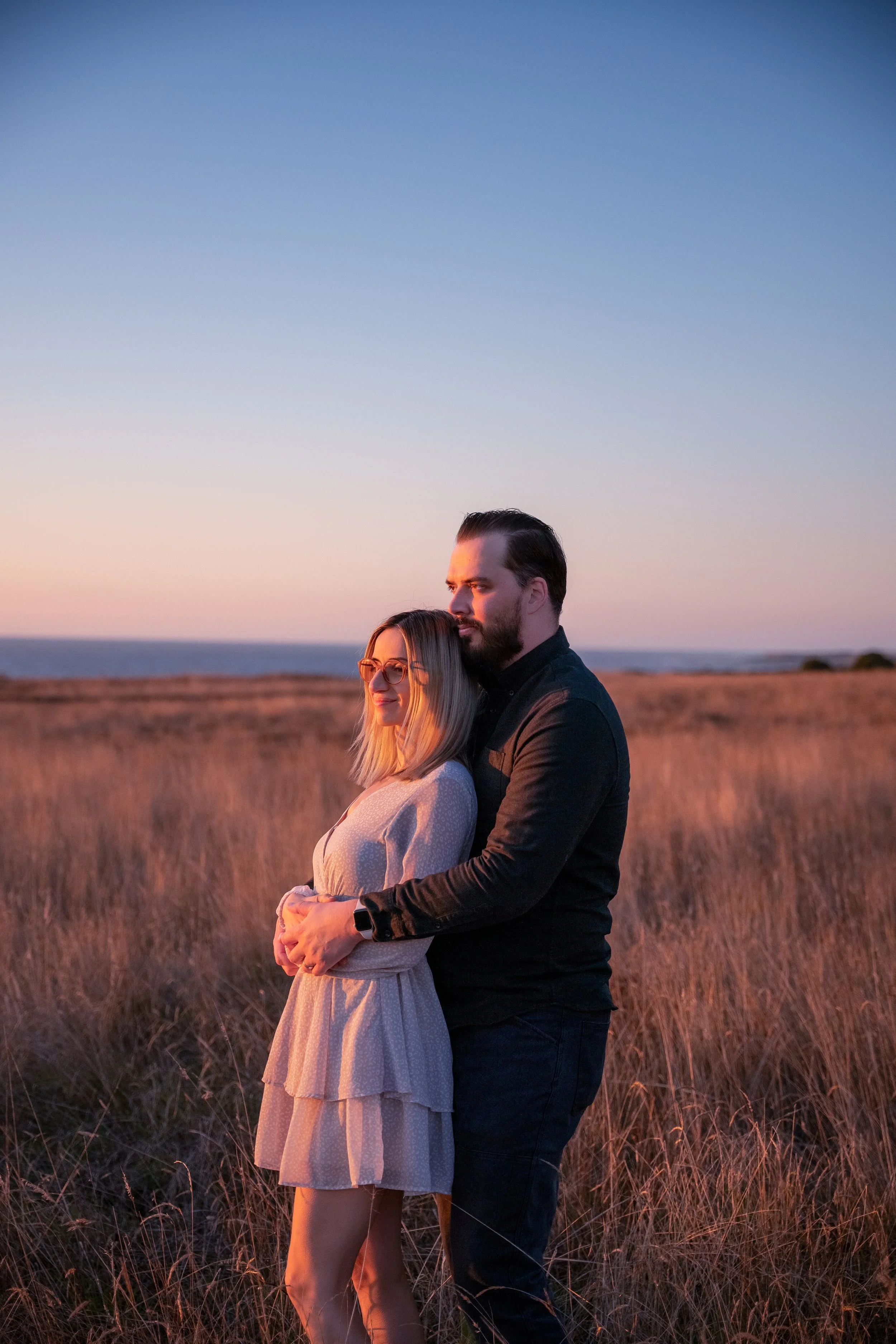 A couple stands close together in a field during sunset, with the woman smiling and the man looking off into the distance.