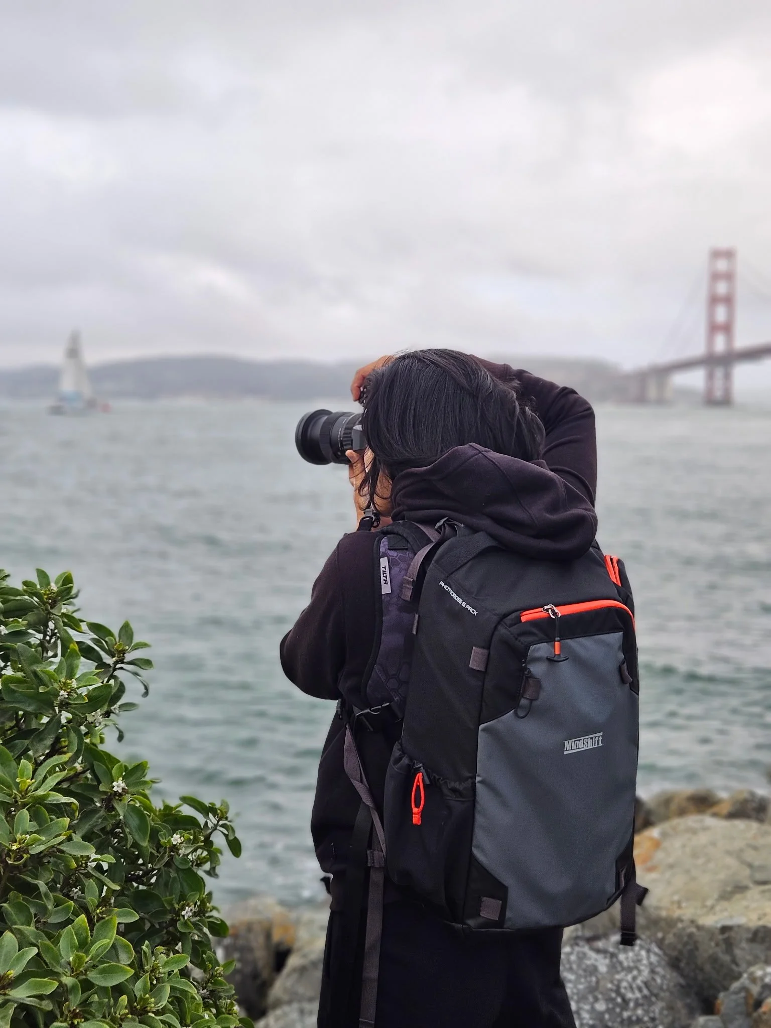 Person with long dark hair taking a photo with a camera near the water, with the Golden Gate Bridge in the background on a cloudy day.