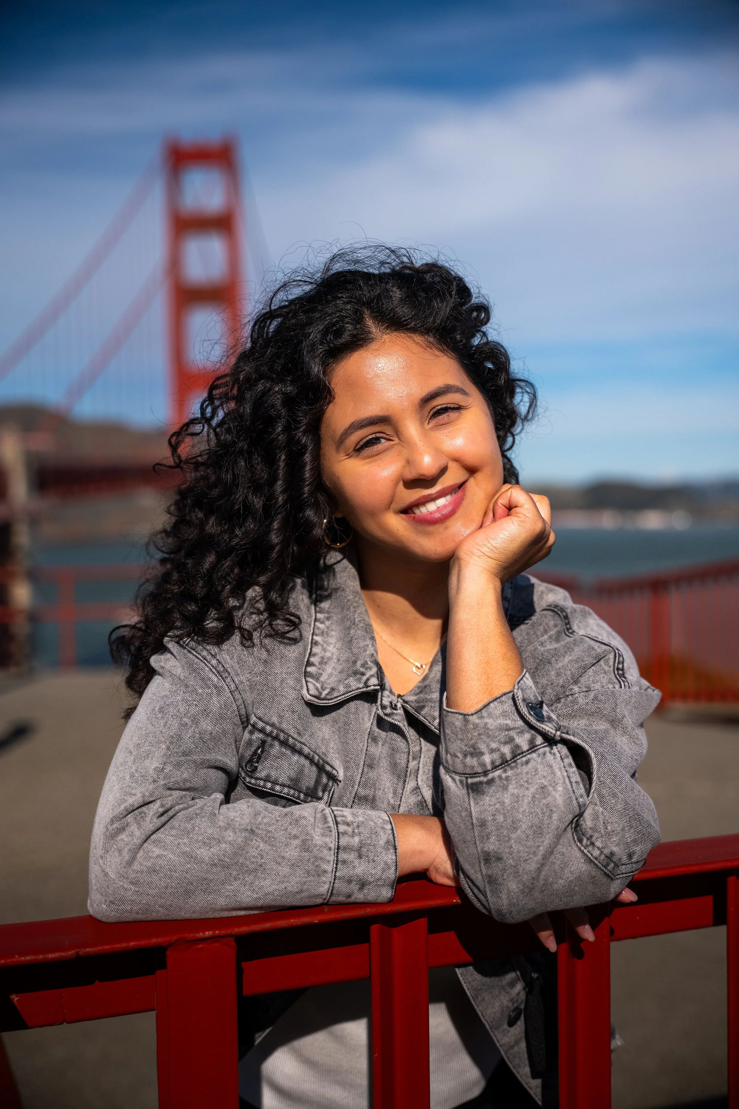 Young woman with curly black hair smiling, resting her chin on her hand, standing near a red railing with the Golden Gate Bridge in the background under a partly cloudy sky.