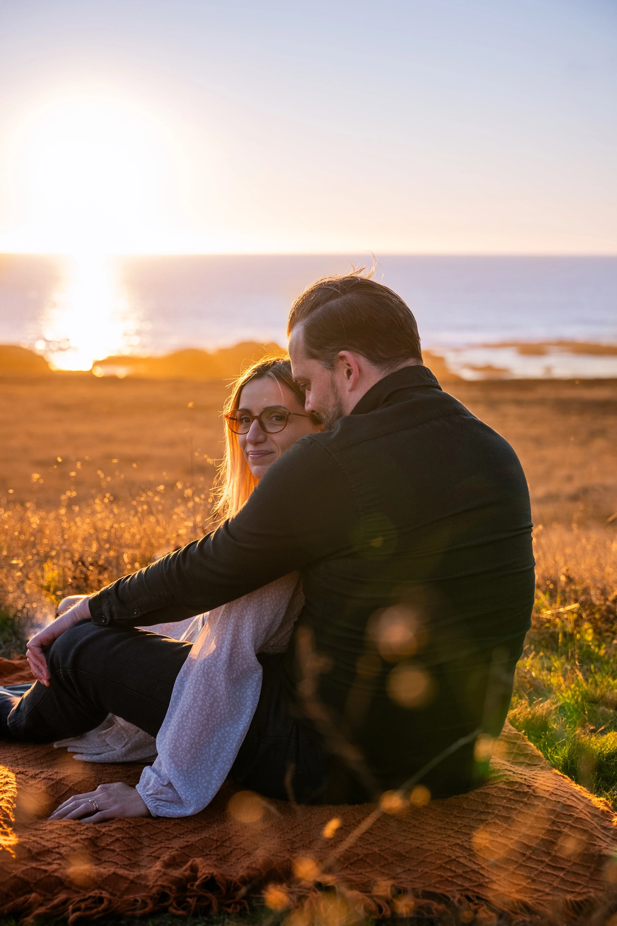 A man and woman sitting on a blanket near the ocean during sunset, sharing an intimate moment with their foreheads touching.