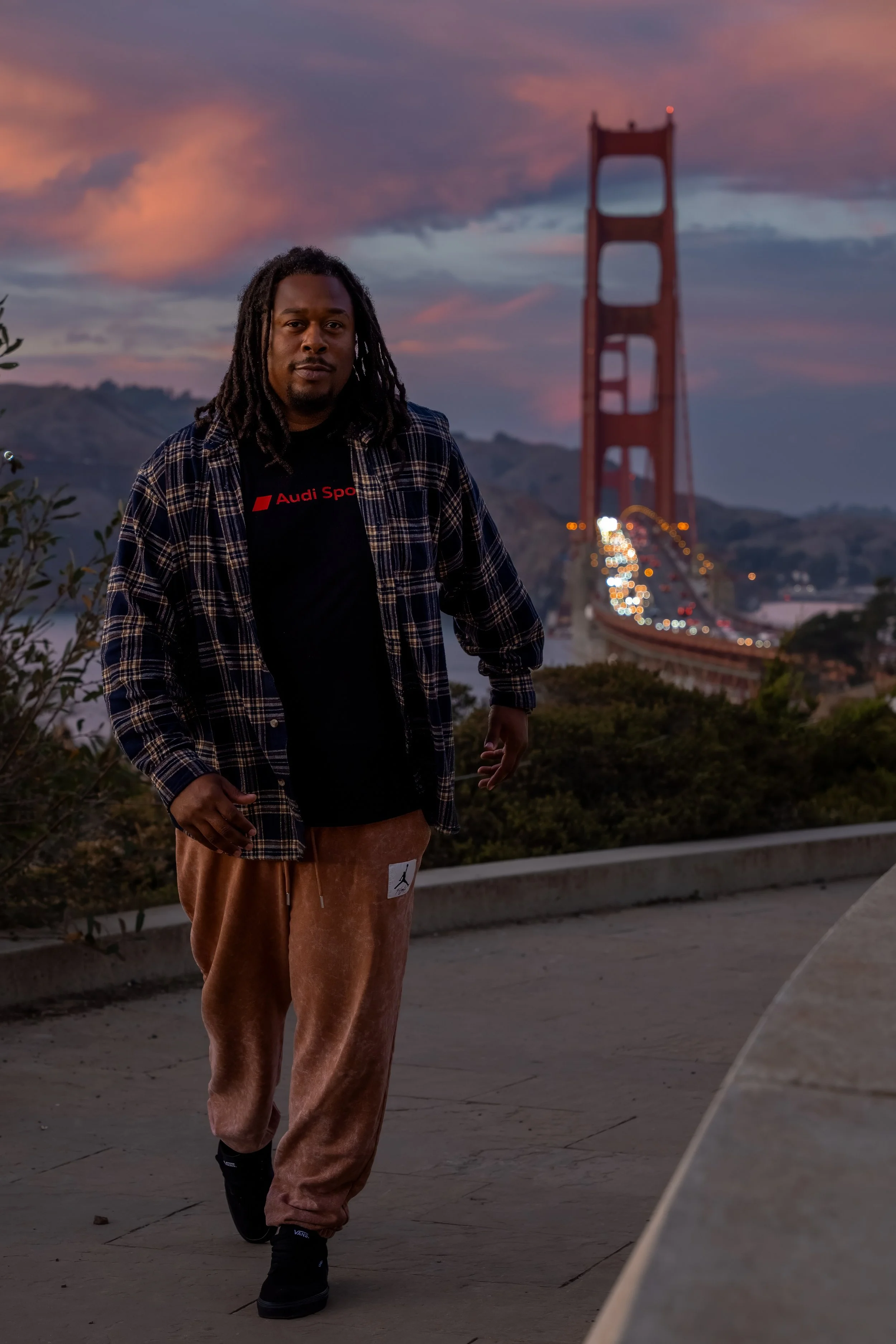 A man with dreadlocks wearing a black t-shirt with red text, an unbuttoned plaid shirt, and brown pants, walking outdoors near the Golden Gate Bridge at sunset.