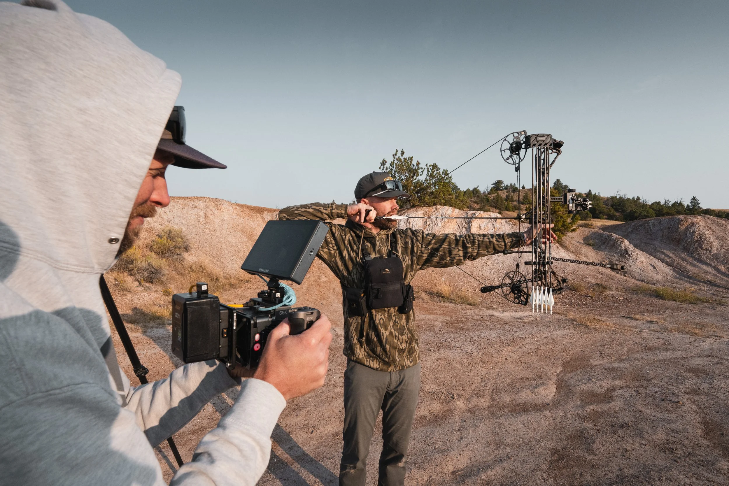 Two guys in an outdoor desert-like area practicing archery with the new Mathews Archery Arc 32, one filming with a freely ember 5k and the other aiming.