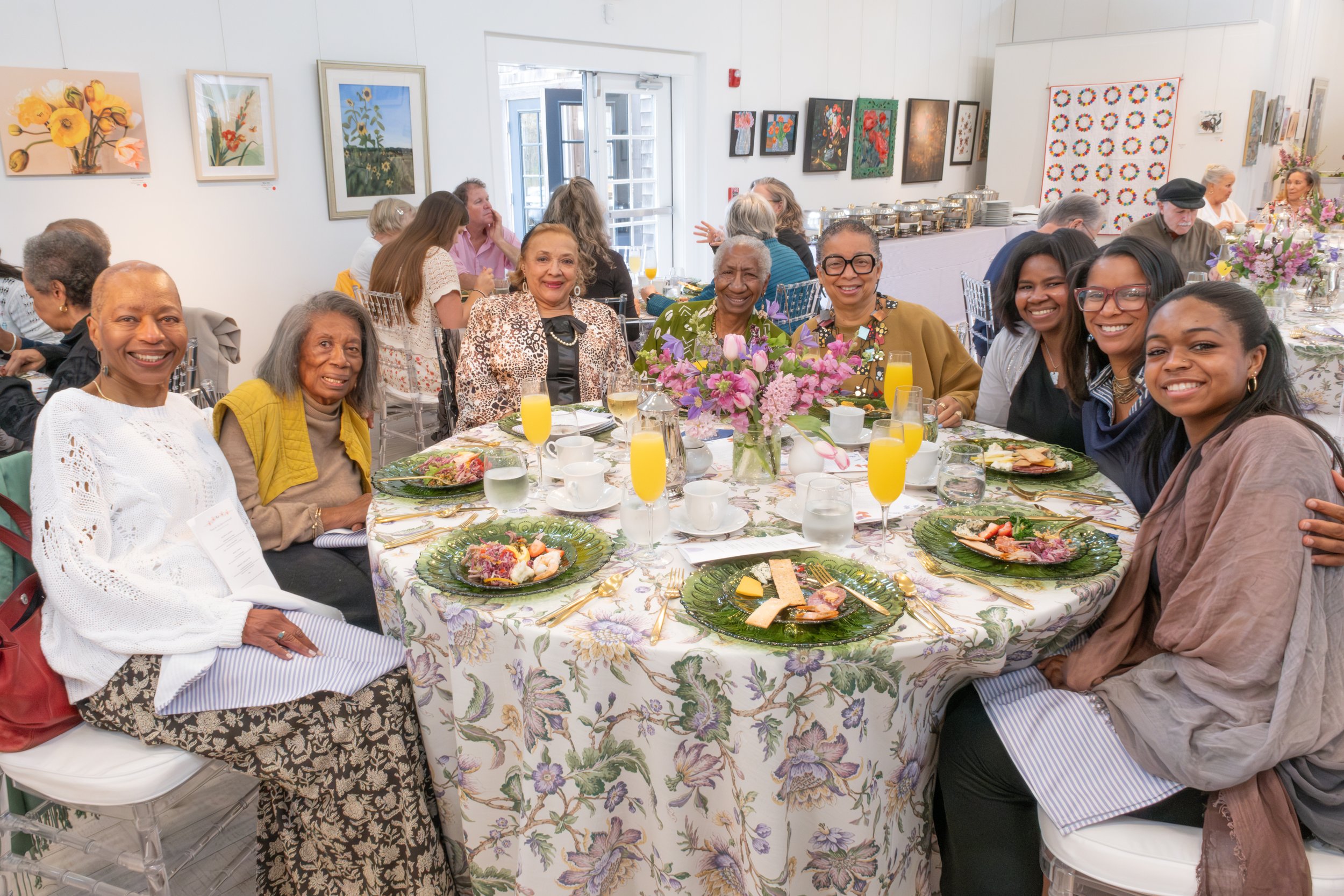 A group of women seated around a dining table at a social gathering or celebration, with art displayed on the walls behind them, and plates of food and drinks on the table.