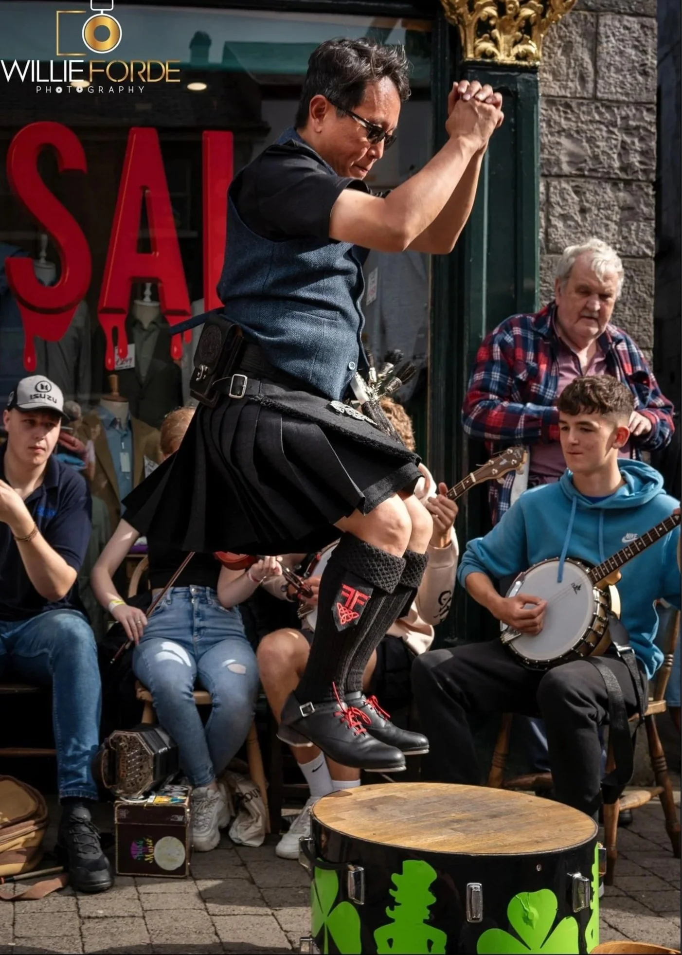 Marcel in a kilt jumping on to a small platform to perform Sean-Nós dance with live street performer band playing in the background, surrounded by onlookers sitting and standing on a sidewalk. Musical instruments include fiddle and banjo.