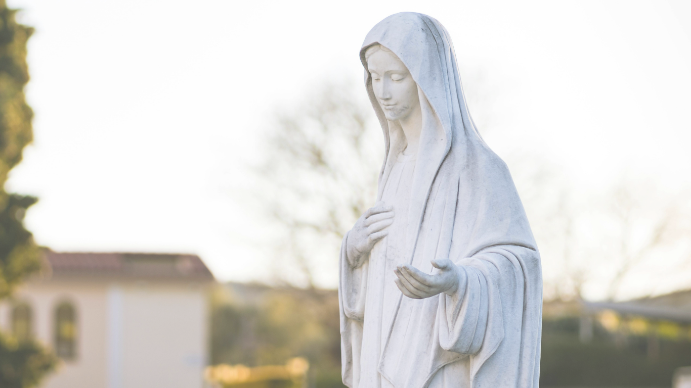 White marble statue of the Virgin Mary with her hands open, looking down with a gentle expression, outdoors with trees and a house in the background.