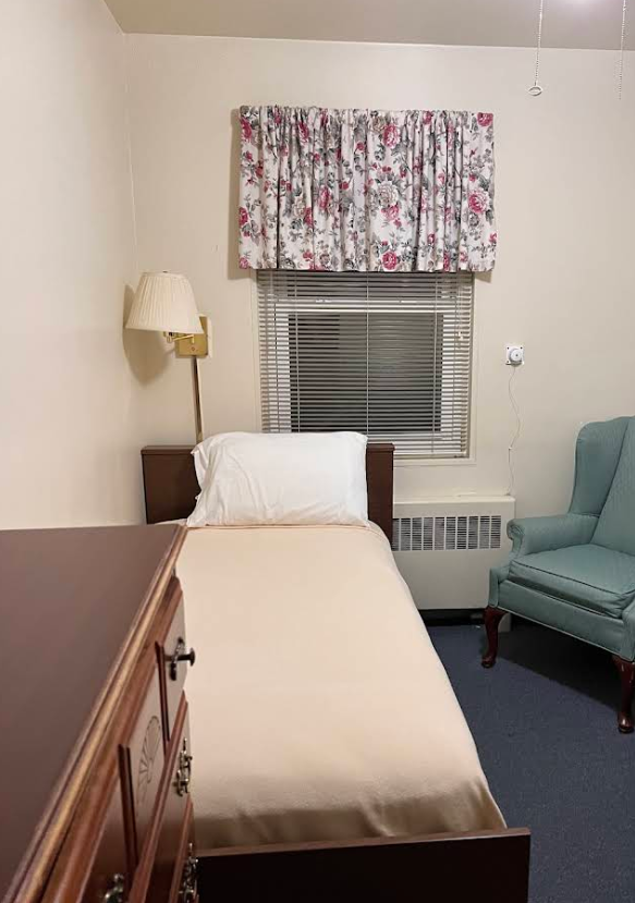A simple hotel room with a single bed, pillow, floral curtains, window with blinds, a wall-mounted lamp, blue armchair, and a wooden dresser.