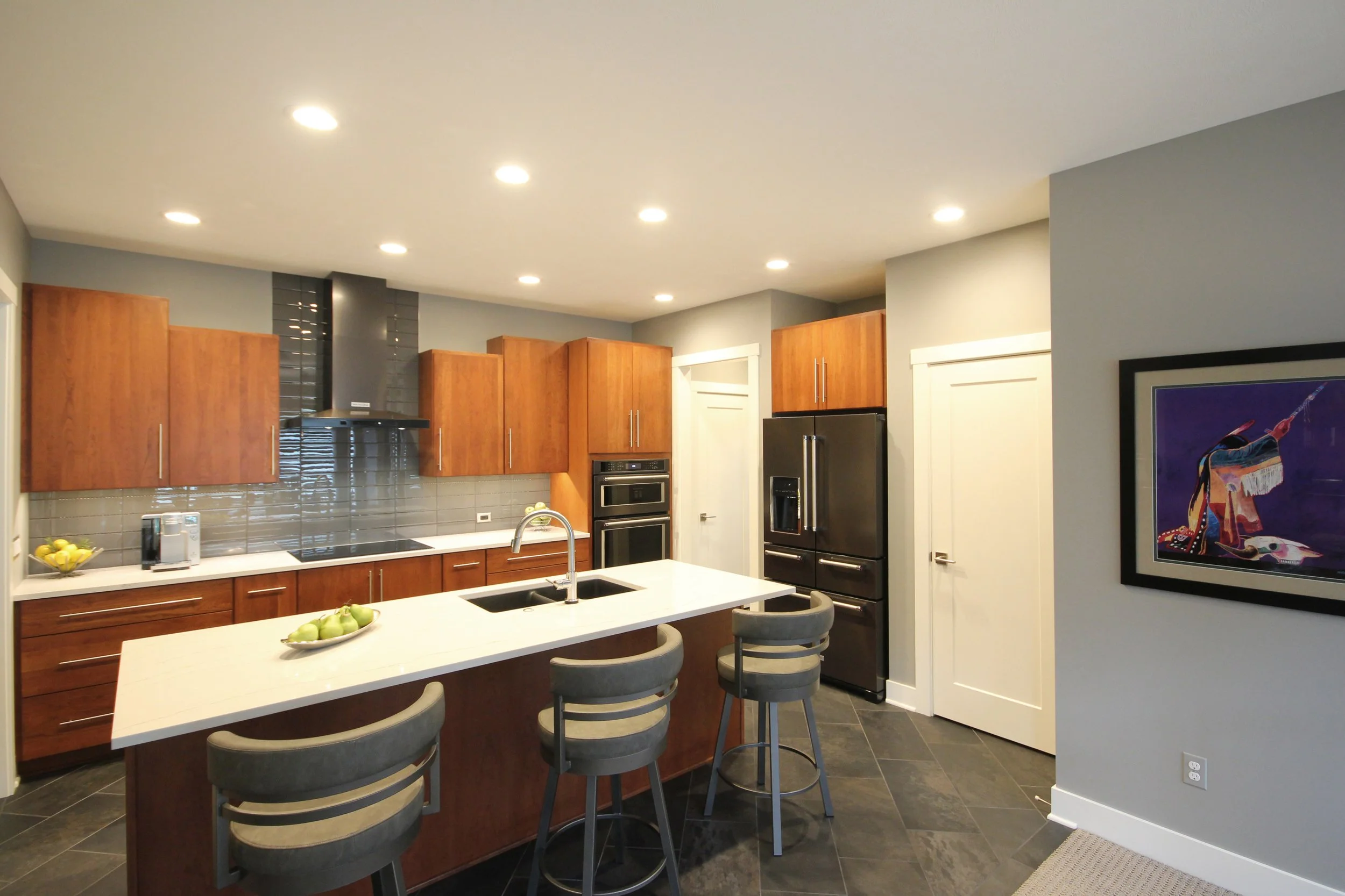 Modern kitchen with wooden cabinets, white countertop island, gray tile backsplash, black appliances, and gray chairs around the island. Recessed ceiling lights and framed artwork on the wall.