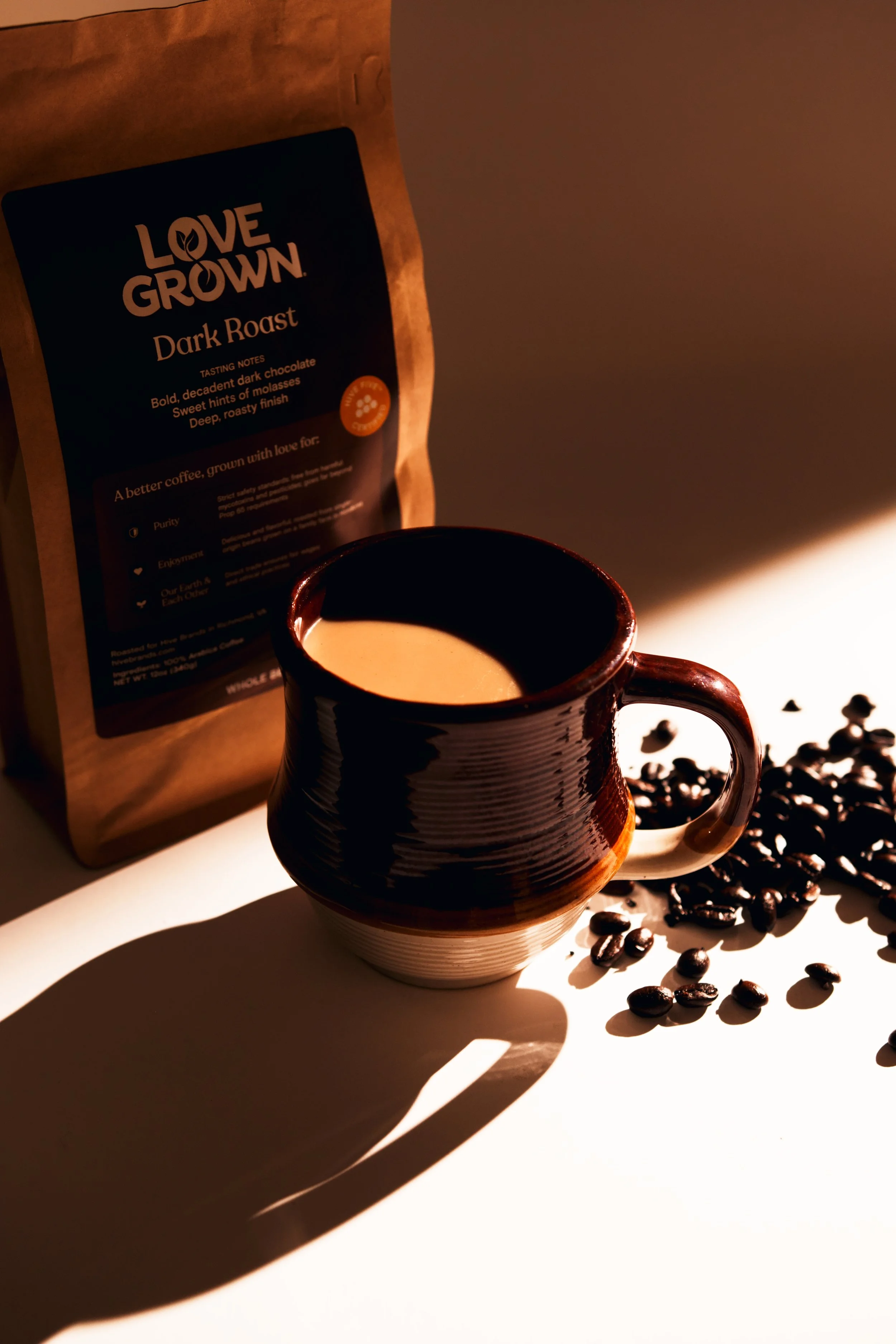 A dark brown coffee mug filled with coffee, with spilled coffee beans scattered next to it, and a paper bag of Love Grown dark roast coffee in the background, placed on a white surface with shadows.