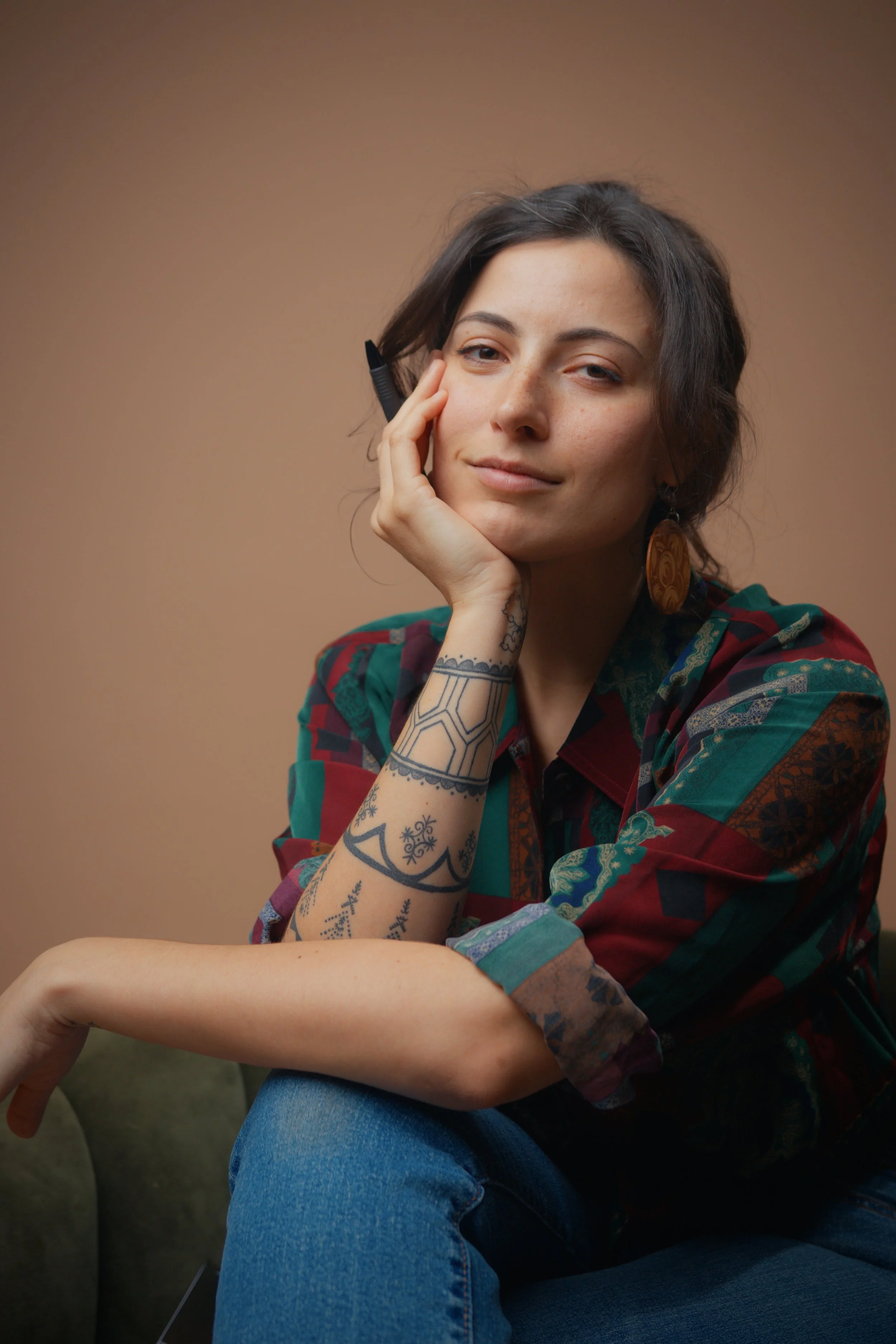 A young woman with tattoos on her arm, wearing a colorful patterned shirt, sitting on a green armchair against a neutral brown background, resting her head on her hand and gazing at the camera.