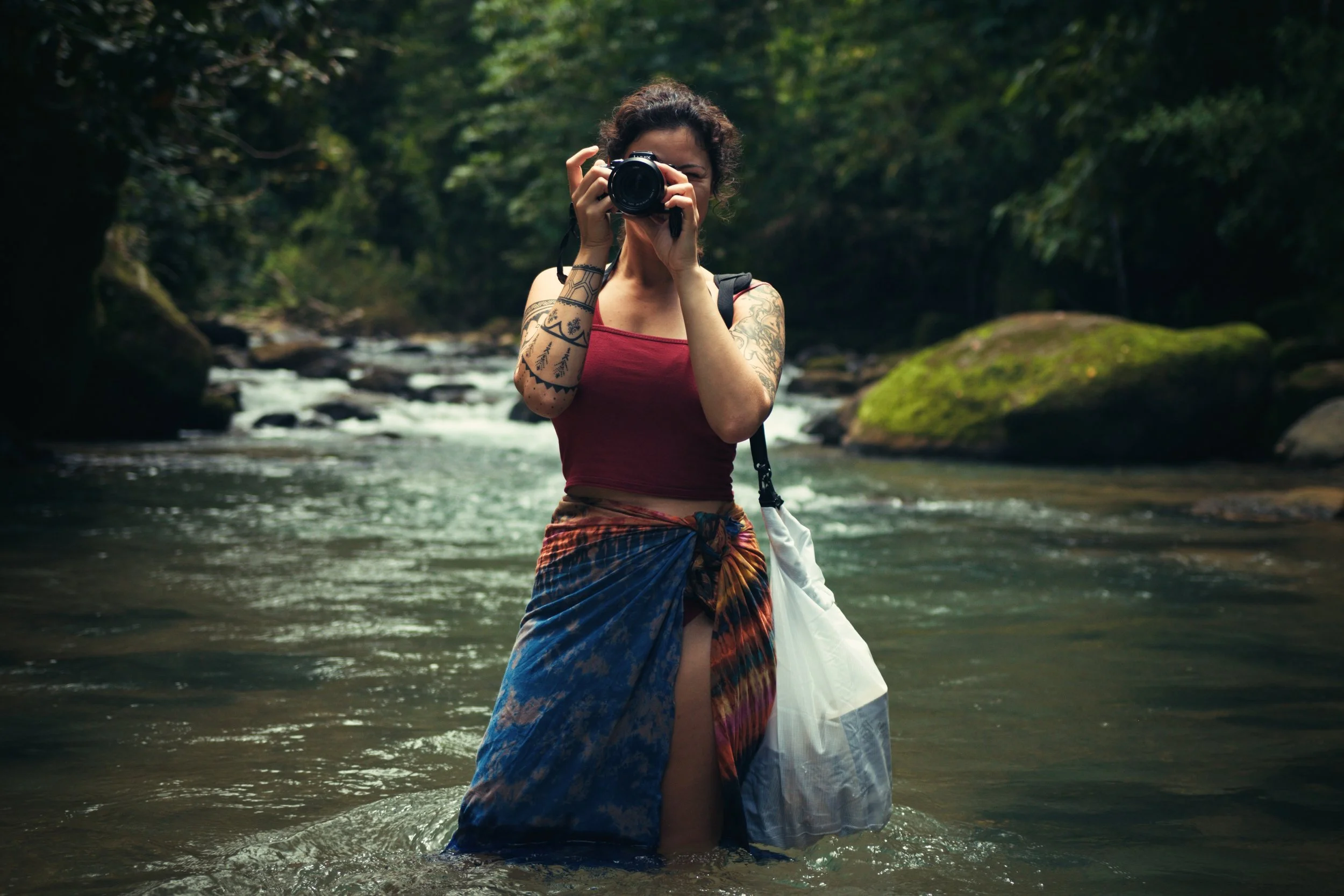 Woman standing in a river, taking a photo with a camera, surrounded by trees and rocks.
