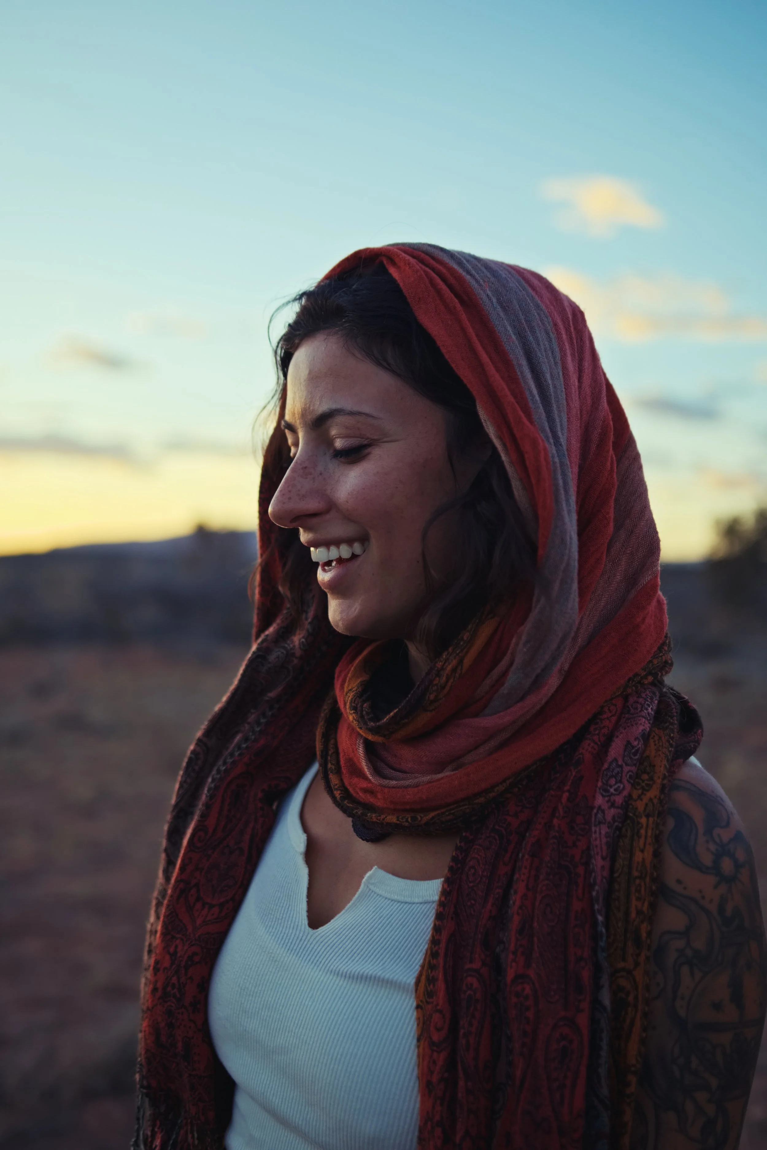 A woman with dark hair wrapped in a red and multicolored scarf, smiling with her eyes closed, outdoors during sunset.