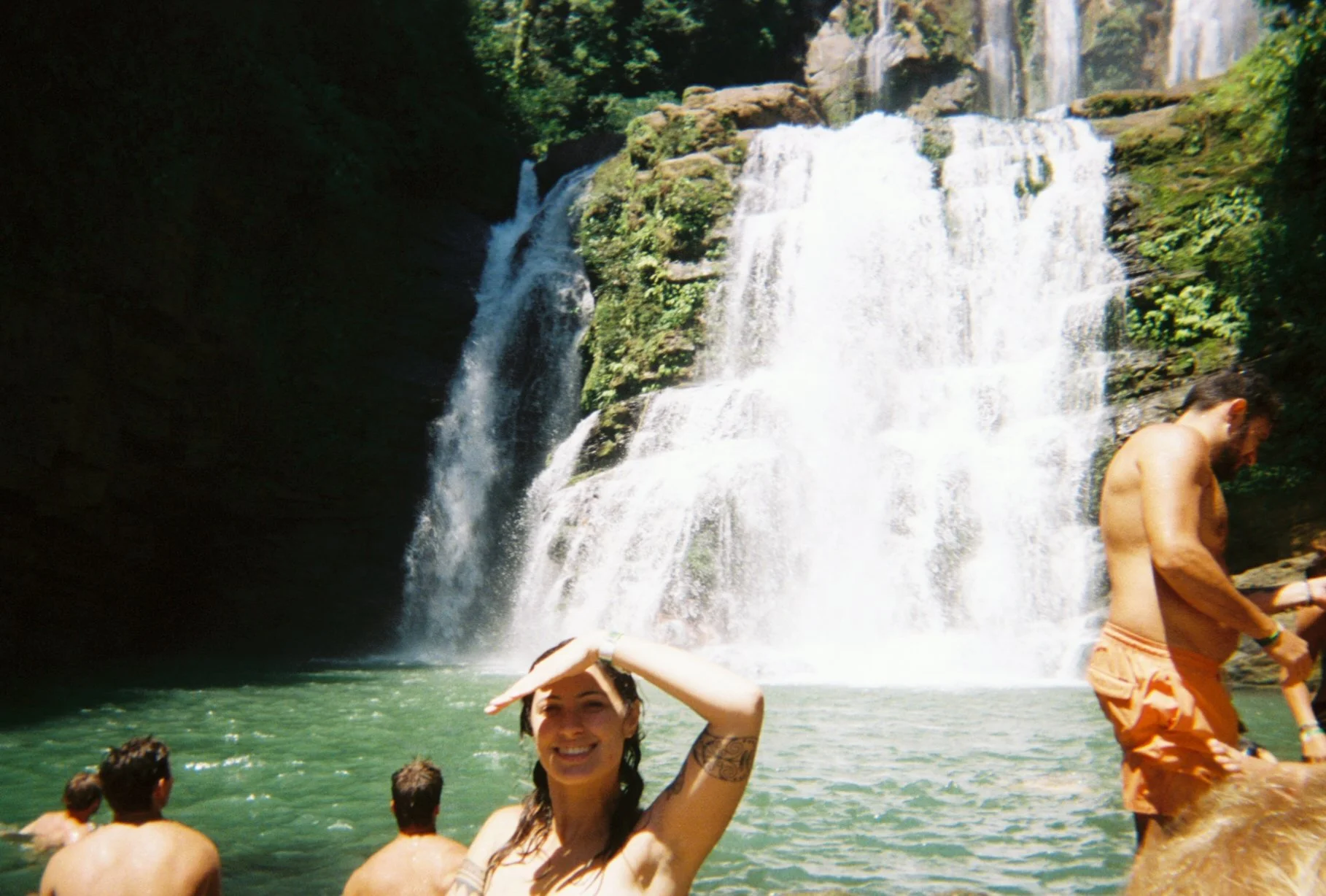 People enjoying swimming in a river near a large waterfall with lush green trees.