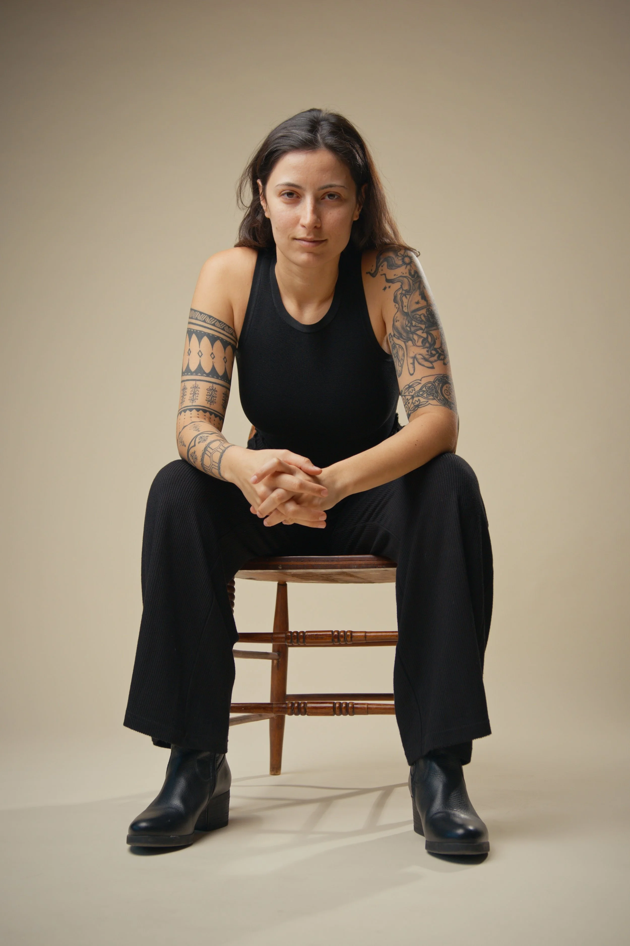 A woman with tattoos on her arms, wearing a black sleeveless top and black pants, sitting on a wooden chair against a plain beige background.