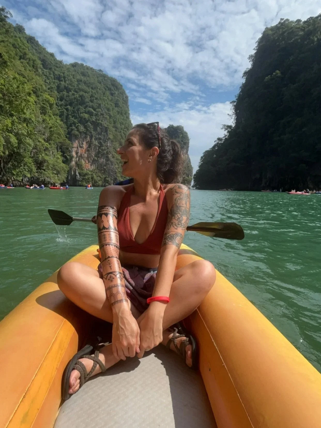 A woman sitting cross-legged on an inflatable yellow kayak, paddling through a river flanked by tall, green, forested cliffs. She is smiling and looking to her left, wearing a red tank top, shorts, and sandals, with tattoos on her arms. The sky above is partly cloudy.