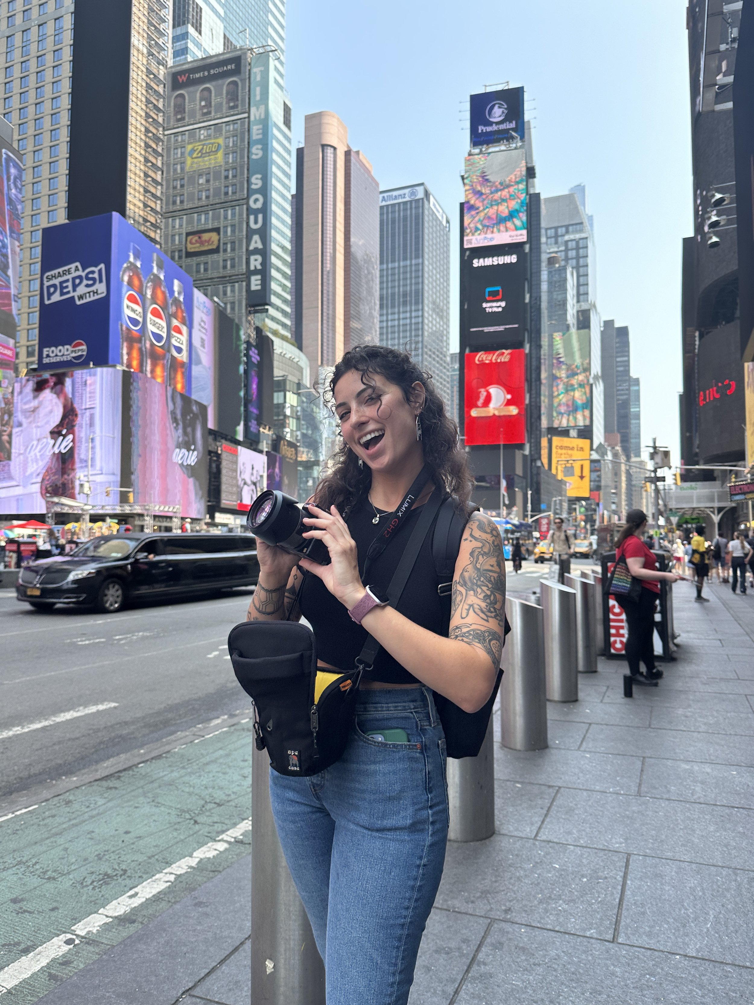 A young woman with curly brown hair, tattoos on her arms, wearing a black top, blue jeans, and a camera around her neck, is smiling and holding a camera in Times Square, New York City, surrounded by tall buildings and digital billboards.