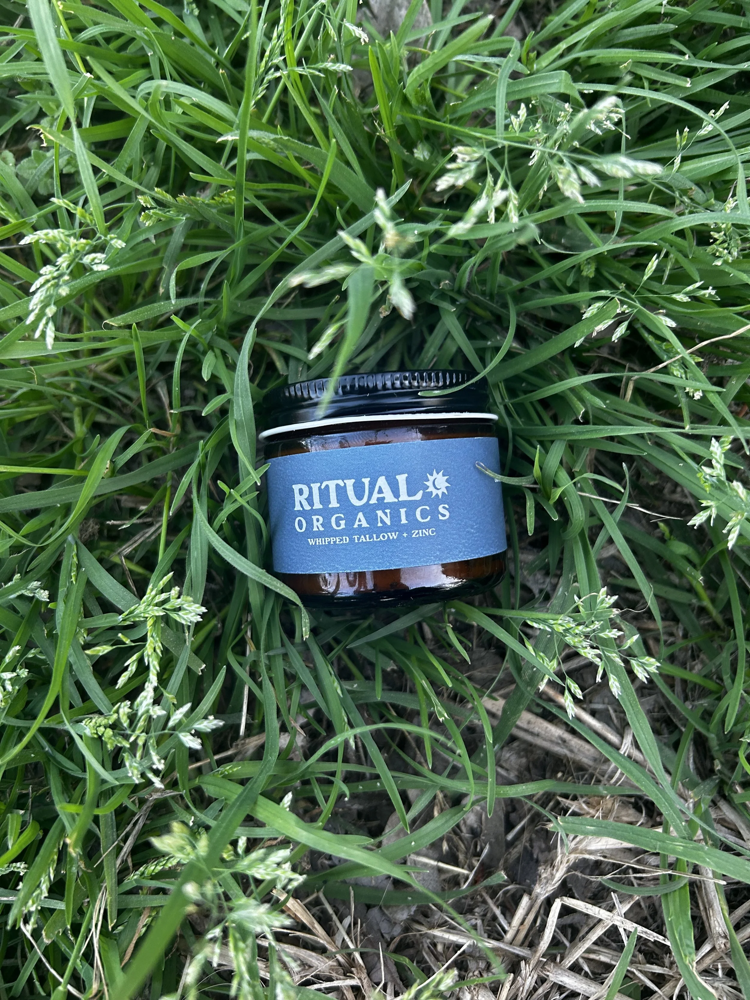 A small jar labeled Ritual Organics, placed on green grass with some small white flowers and blades of grass around it.