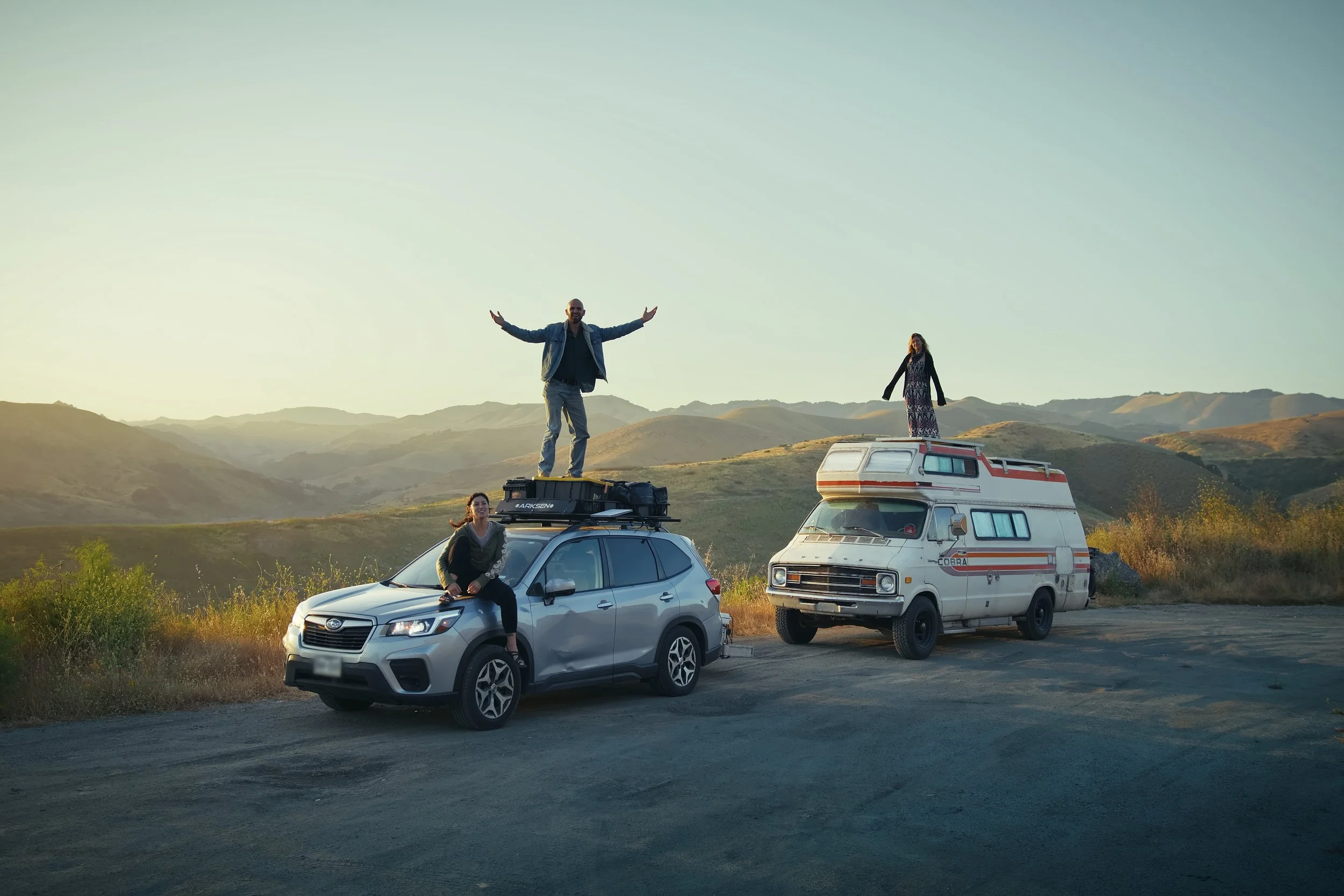 Four people enjoying a scenic mountain view with a car and a camper van, some standing on top of vehicles and others sitting, during sunset.